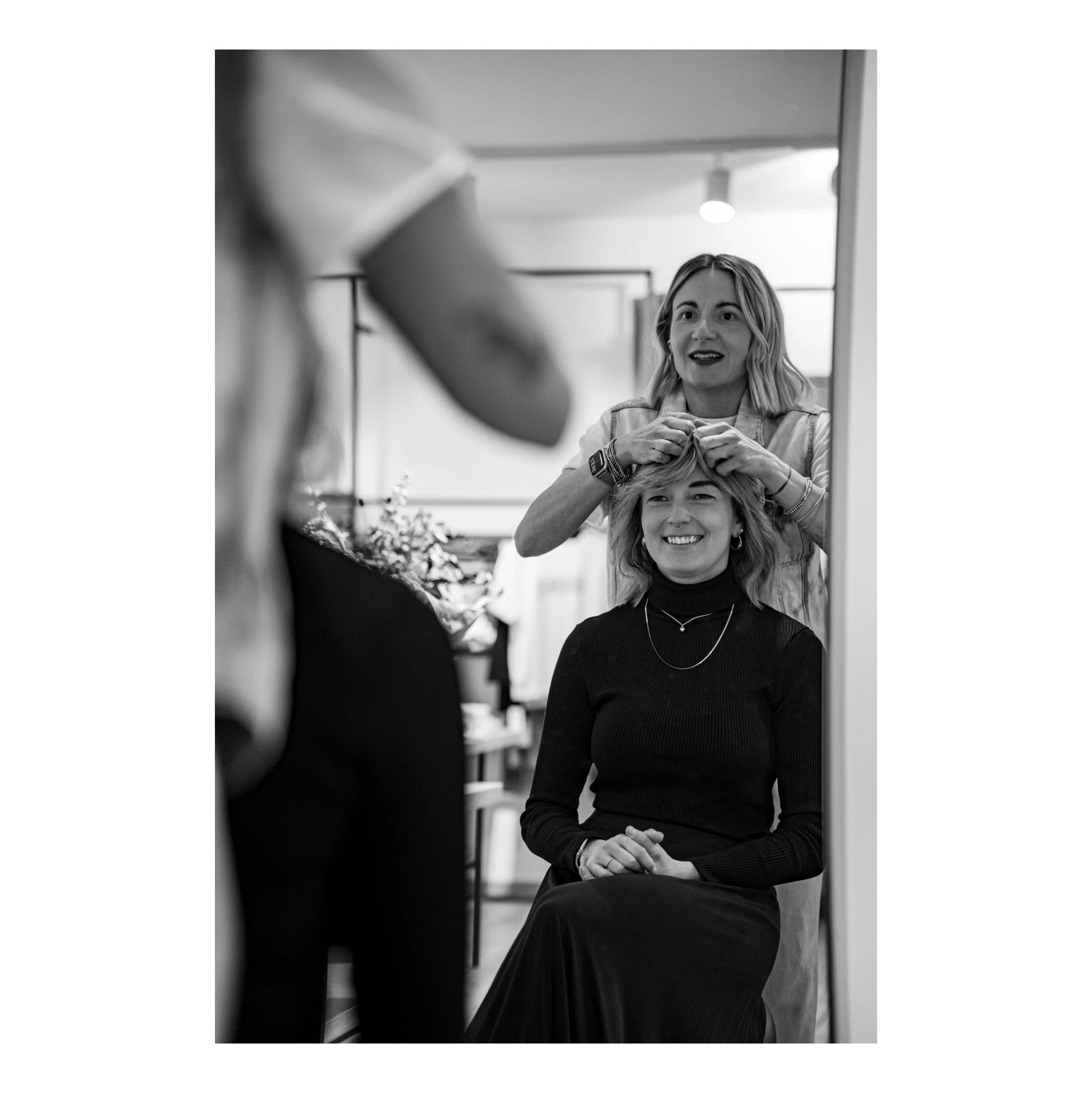 Shooting Business Reportage Friseur/Modehaus. Junge Frau mit braunen Haaren sitzt in einem Raum, während eine andere Person hinter einem Spiegel an ihrem Haar arbeitet. Die Szene ist schwarz-weiß.