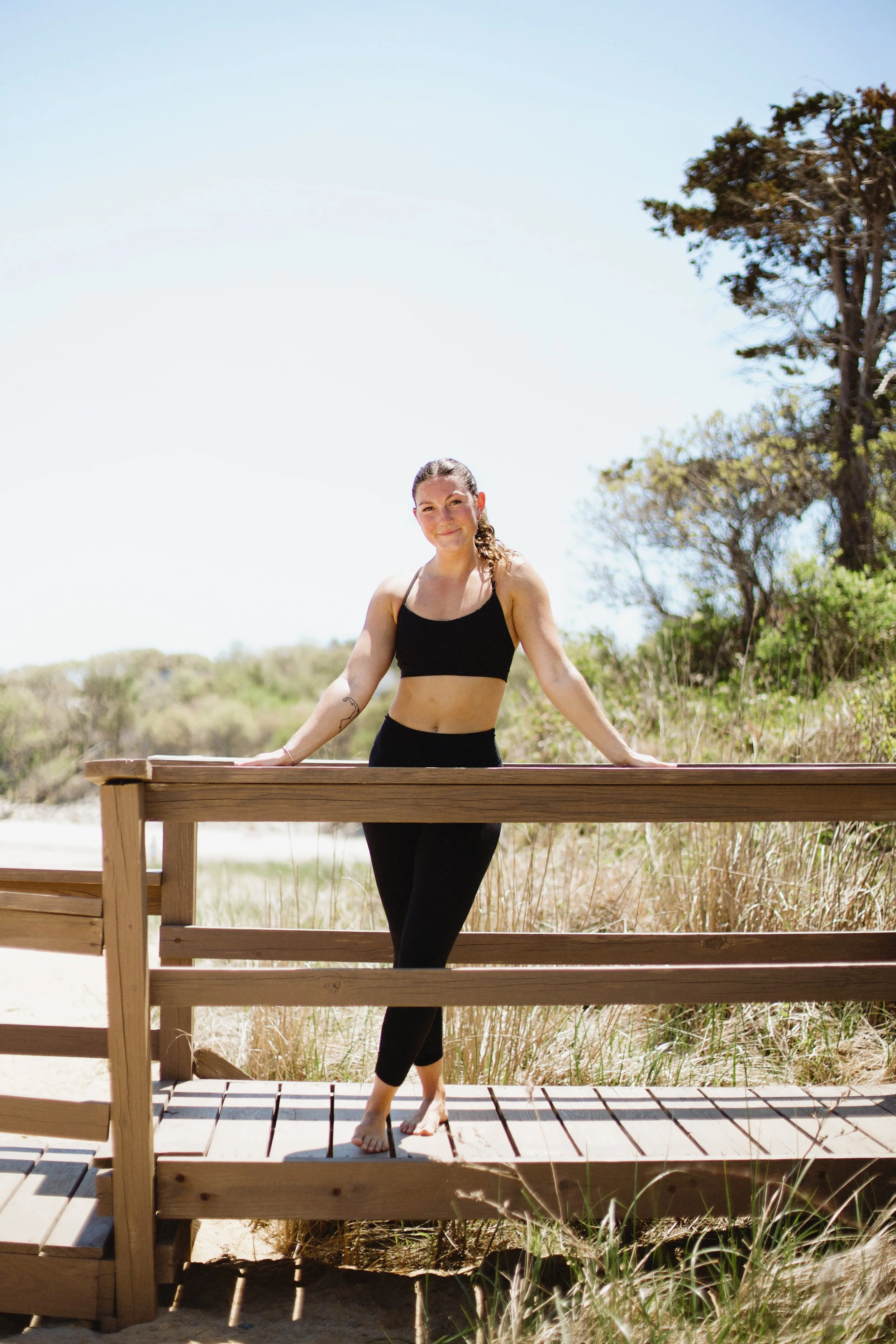Young woman standing on a wooden boardwalk outdoors, smiling, with trees and bushes in the background.