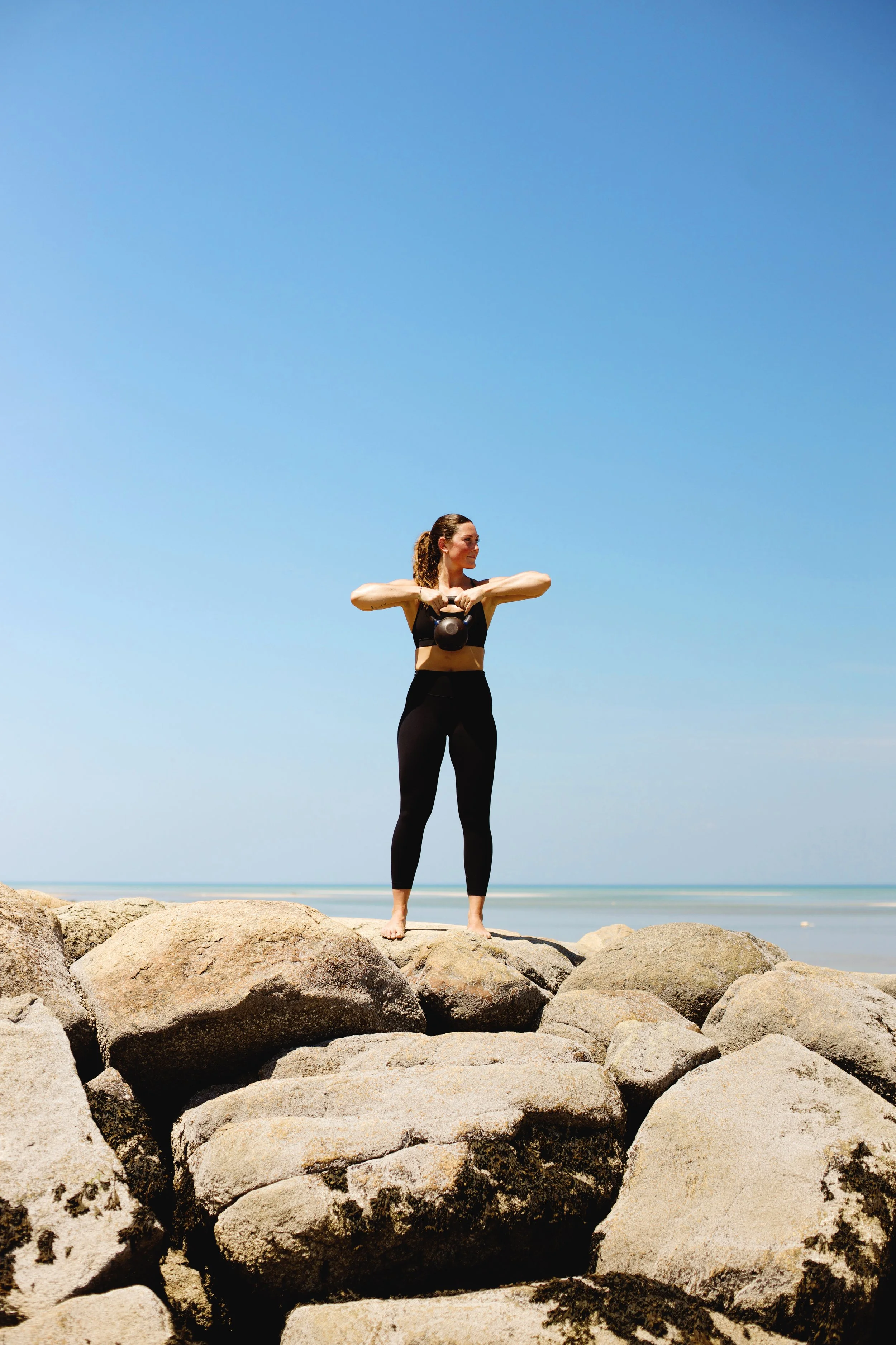 A woman in black workout clothes standing barefoot on rocks by the beach, holding a kettlebell with both hands, looking to her left, with a clear blue sky and ocean in the background.