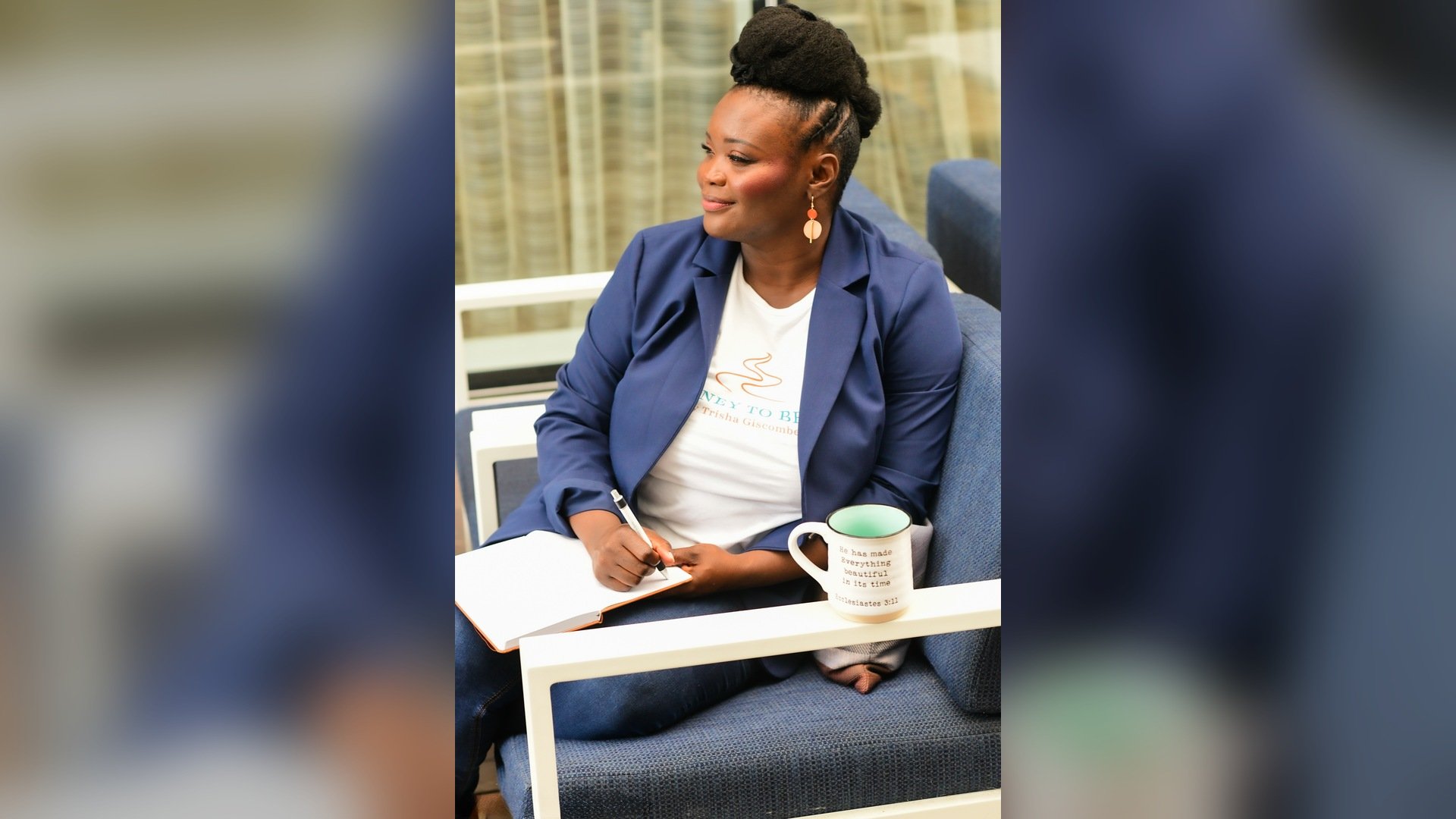 Black woman with natural updo sitting in a blue cushioned chair with a journal and pen in hand looking off to the side in reflection with a mug with a verse sitting on the arm of the chair.