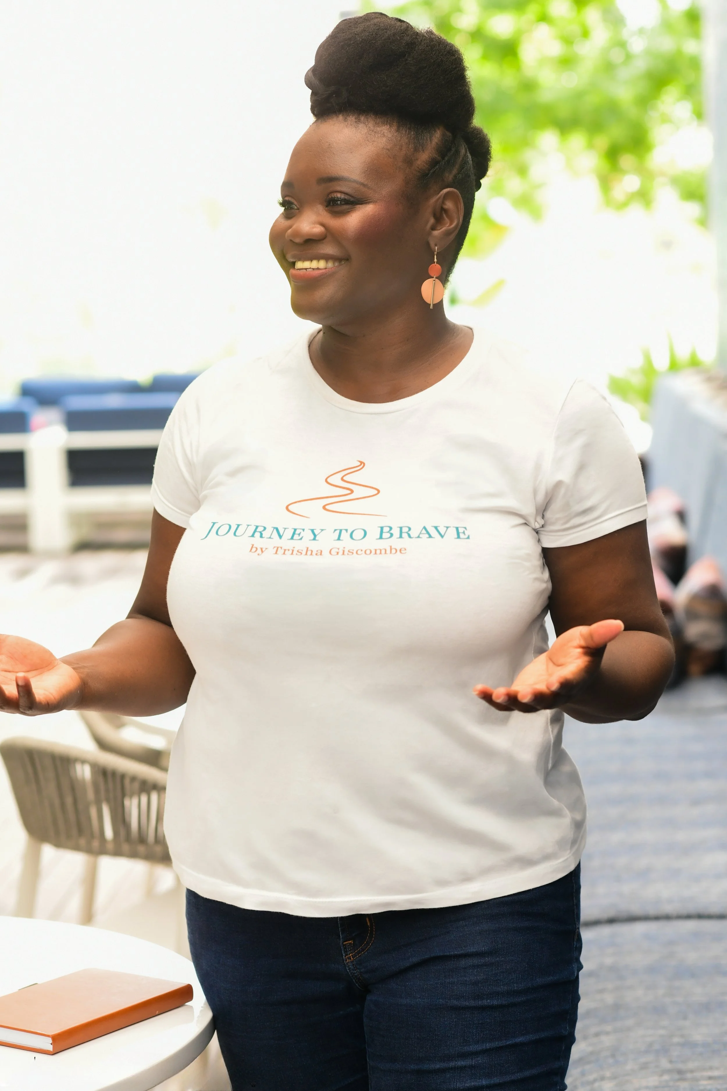 Black woman with a natural updo standing and speaking to an audience with a smile on her face and hands outstretched wearing a white t-shirt with Journey to Brave logo.