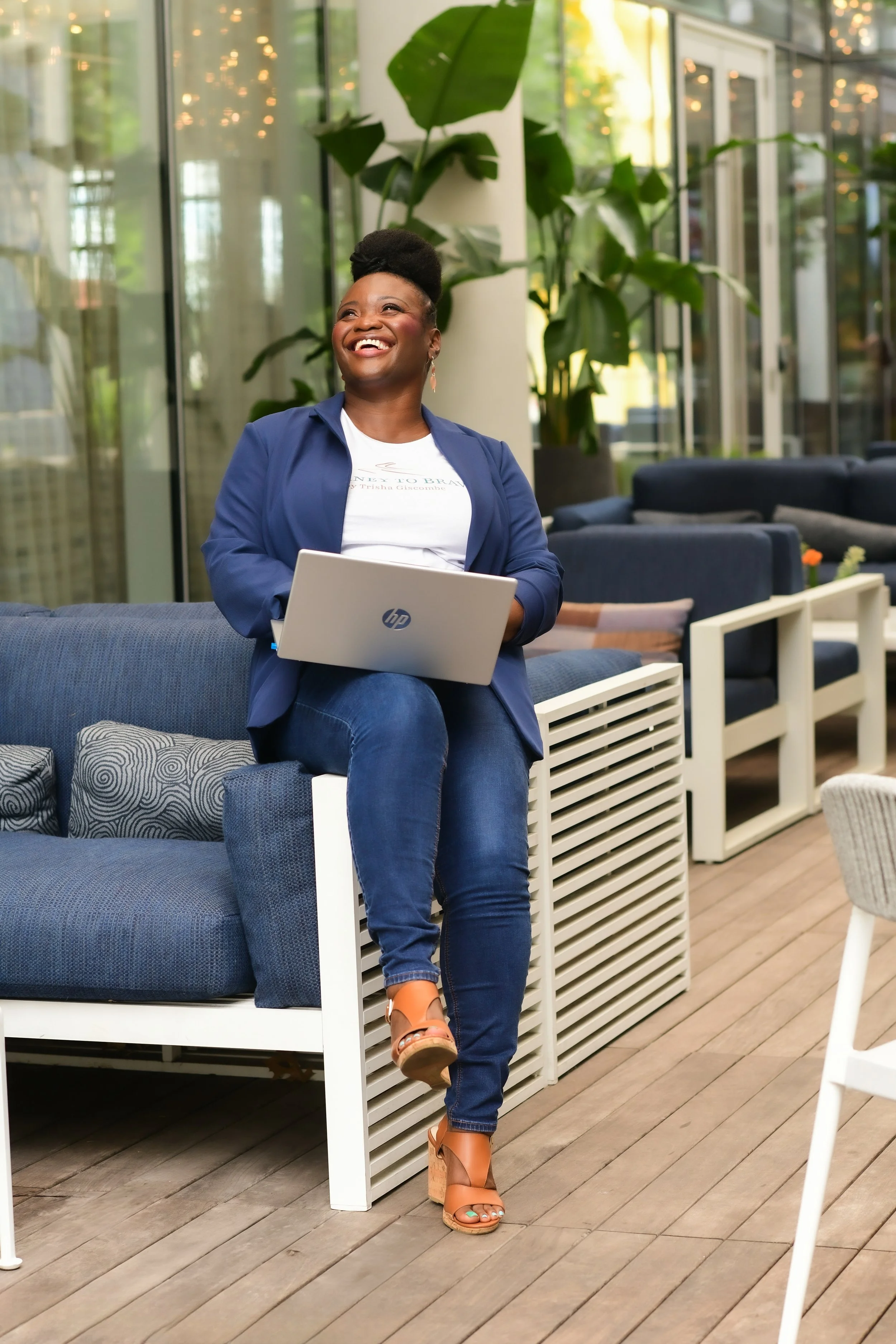Empowered woman full of peace and joy to show healing sitting on the arm of a blue patio sofa, wearing a blue blazer, white t-shirt with Journey to Brave logo, dark blue jeans, and brown wedges with a laptop in hand.