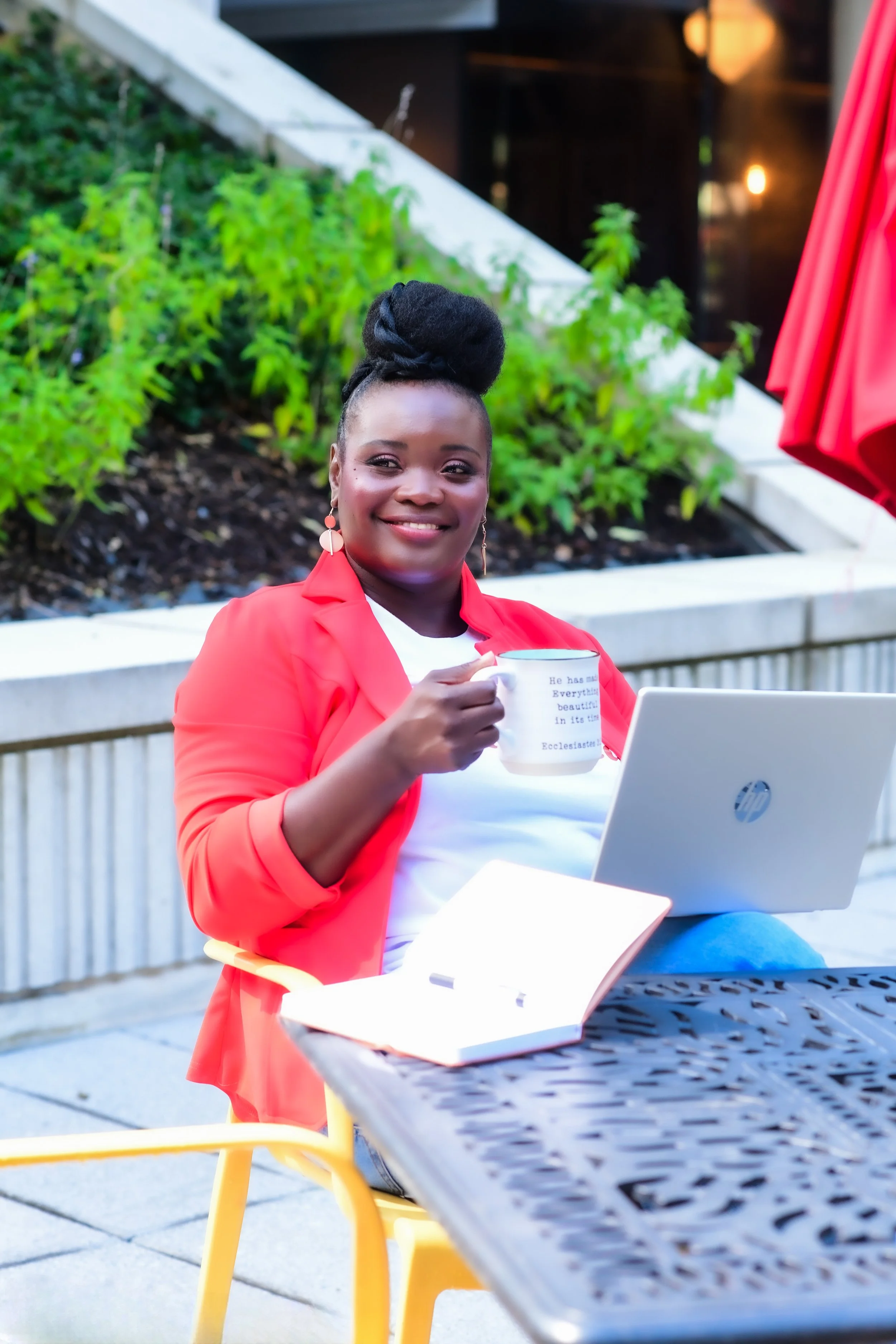 Black woman with natural updo sitting at a black table outside with a mug in hand, journal and pen, and laptop, while looking off to the right reflecting with a smile on her face.