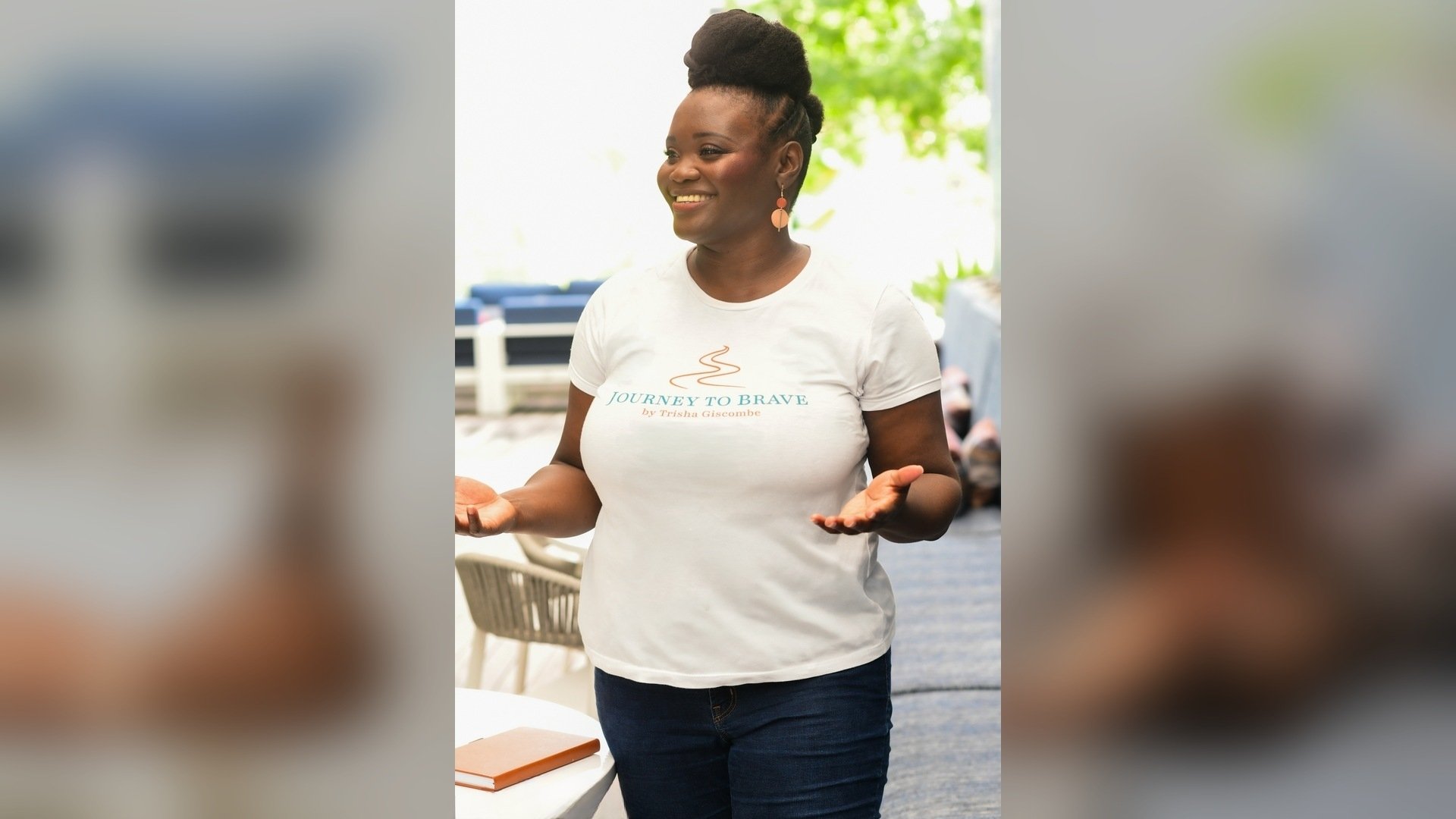 Black woman with a natural updo standing and speaking to an audience wearing a white t-shirt with Journey to Brave logo with her hands outstretched in front of her.
