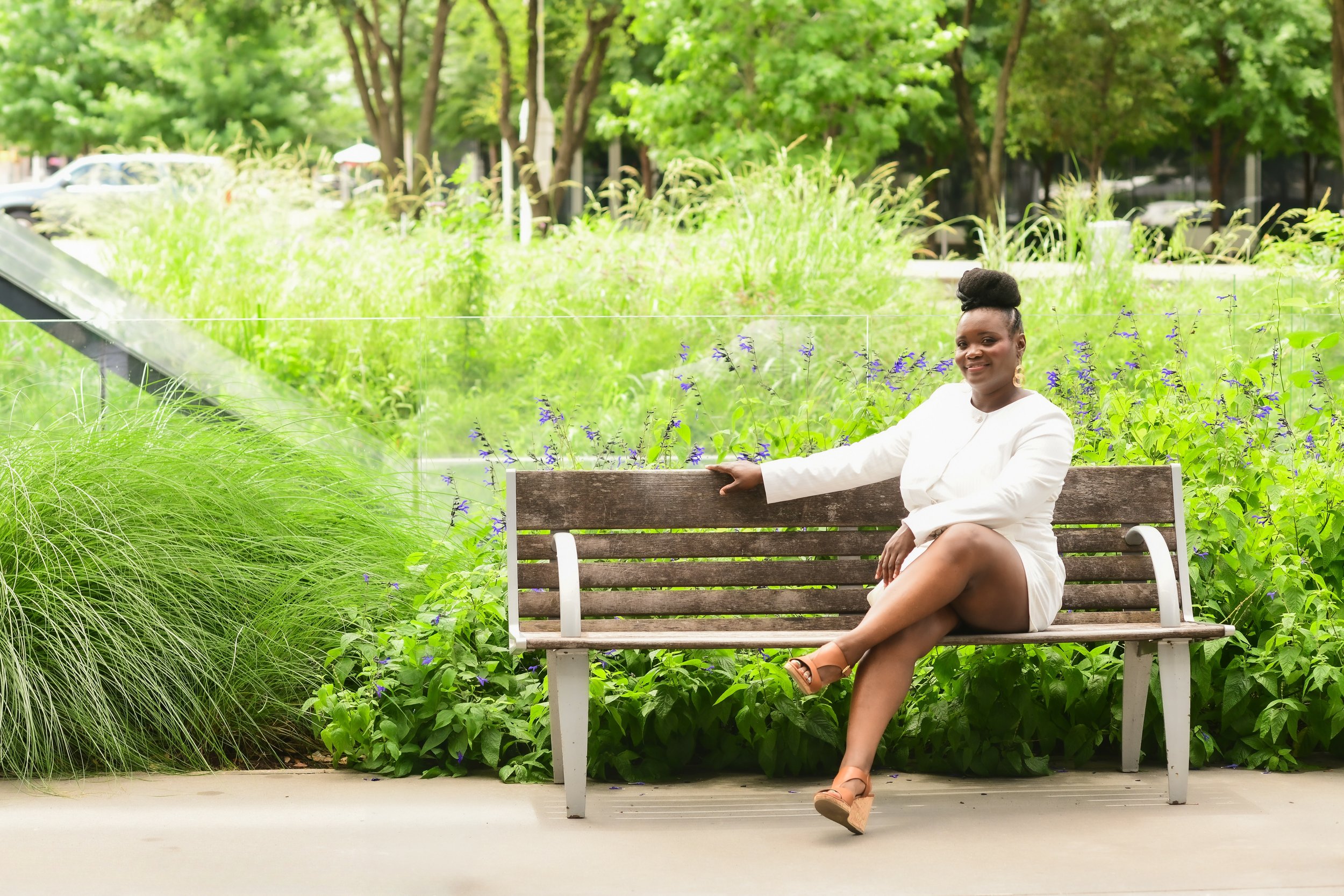 Black woman sitting on a bench looking forward with arm on lap and other hand on the back of the bench and smiling.