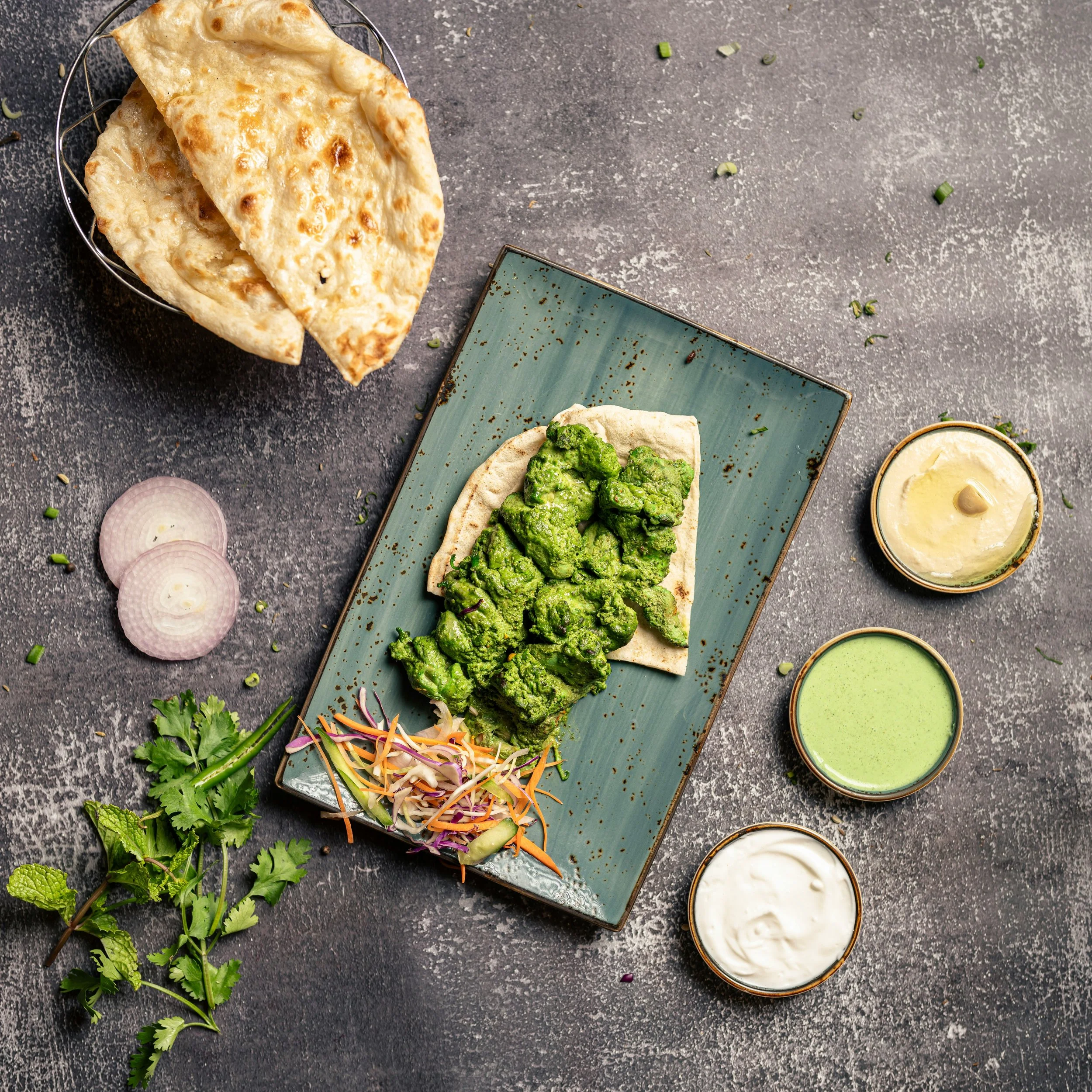 Flat lay of Indian cuisine with naan bread topped with mashed green herbs, small bowls of sauces or dips, and fresh herbs on a dark textured surface.
