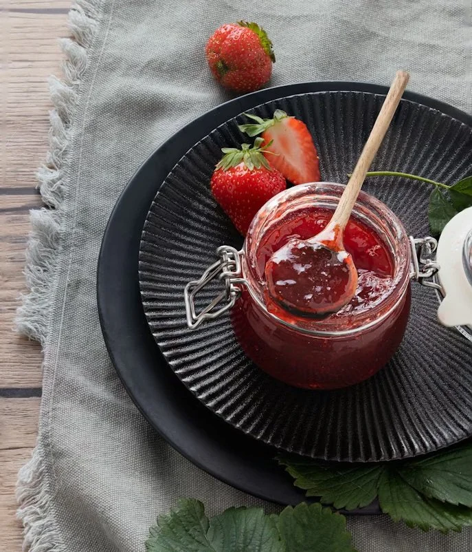 A small glass jar of strawberry jam with a wooden spoon inside, topped with whole and sliced strawberries, on a black textured plate with a larger black plate underneath, on a light gray cloth and wooden surface.