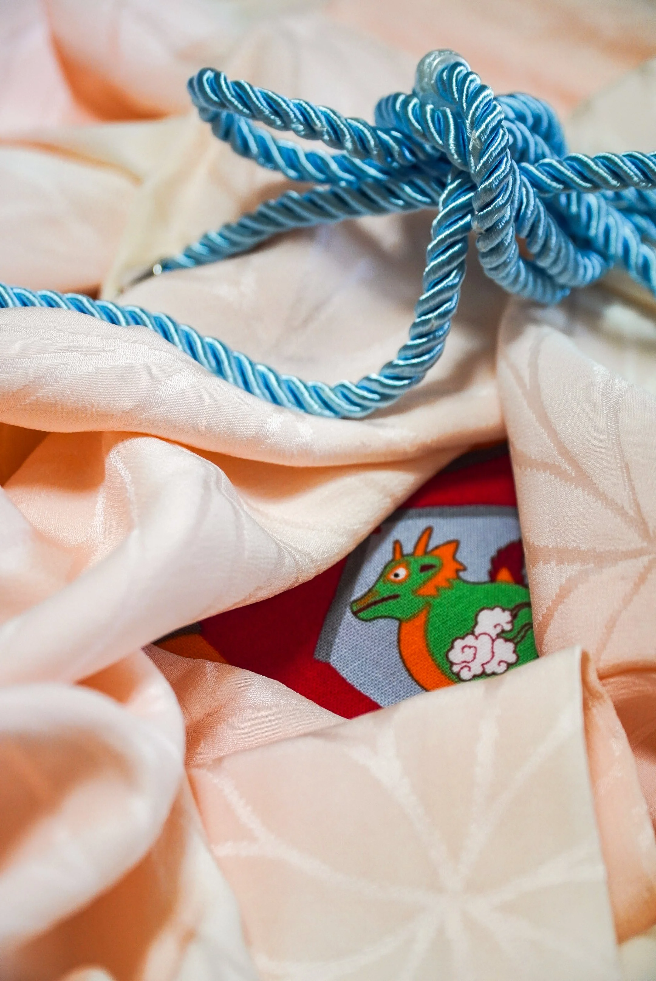 Close-up of a blue twisted cord with a clear plastic tip, lying on soft, pastel-colored fabric with a dragon illustration visible underneath.