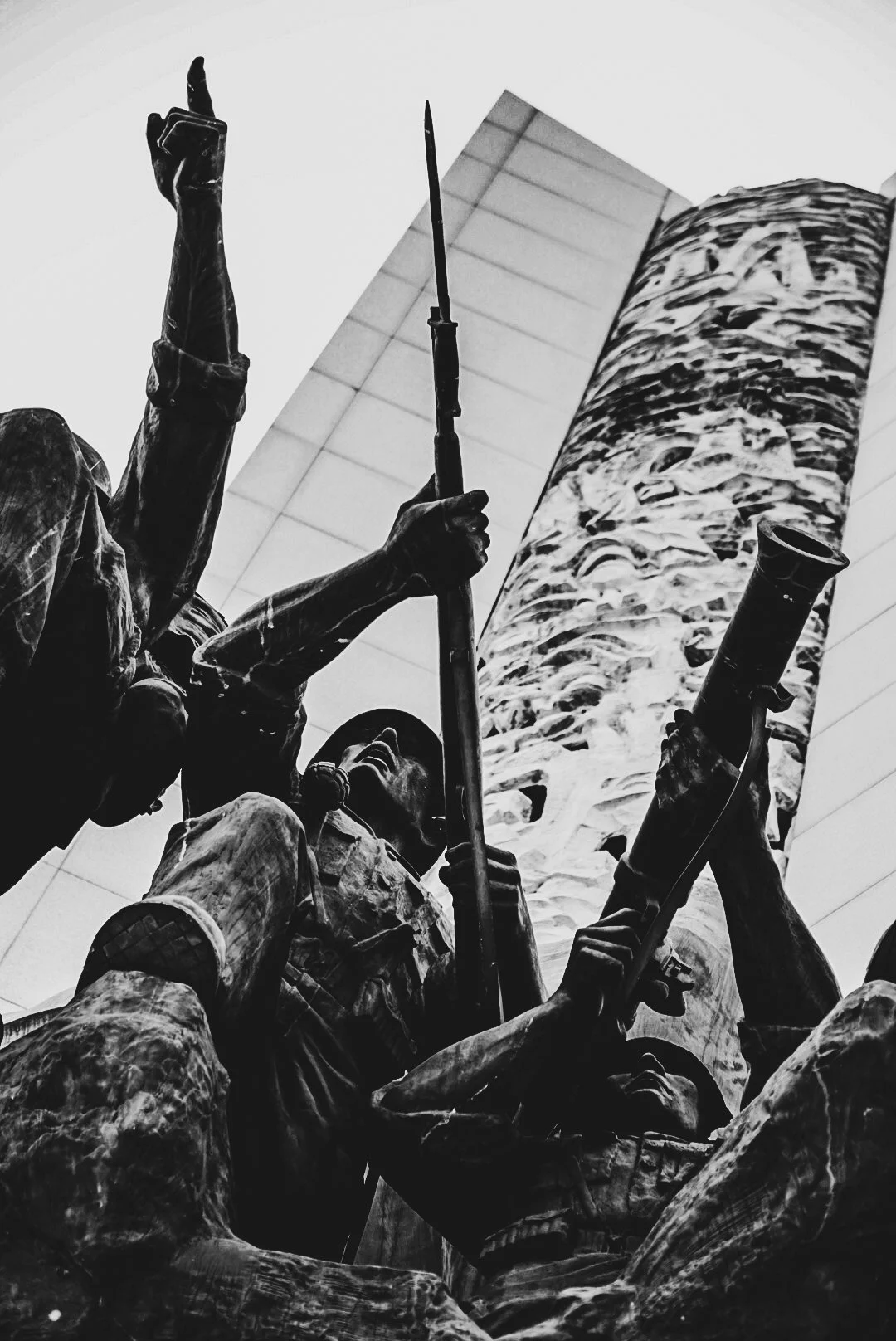 Close-up of a bronze sculpture of soldiers, showing them holding rifles and a telescope, in front of a tall modern building.