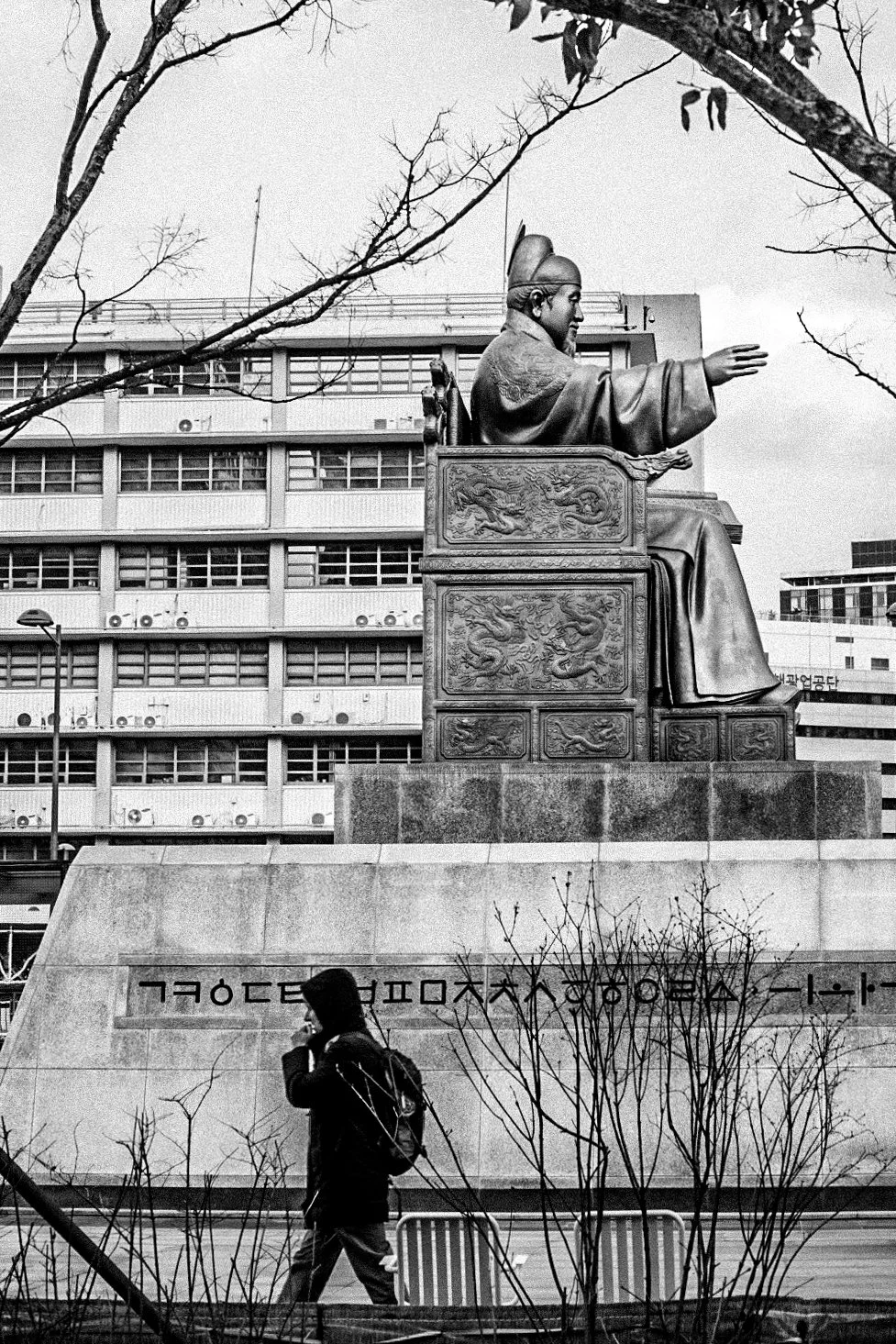 A large statue of a seated historical figure with an extended arm, in front of a modern building. A person with a backpack is walking past in the foreground, with leafless trees around.