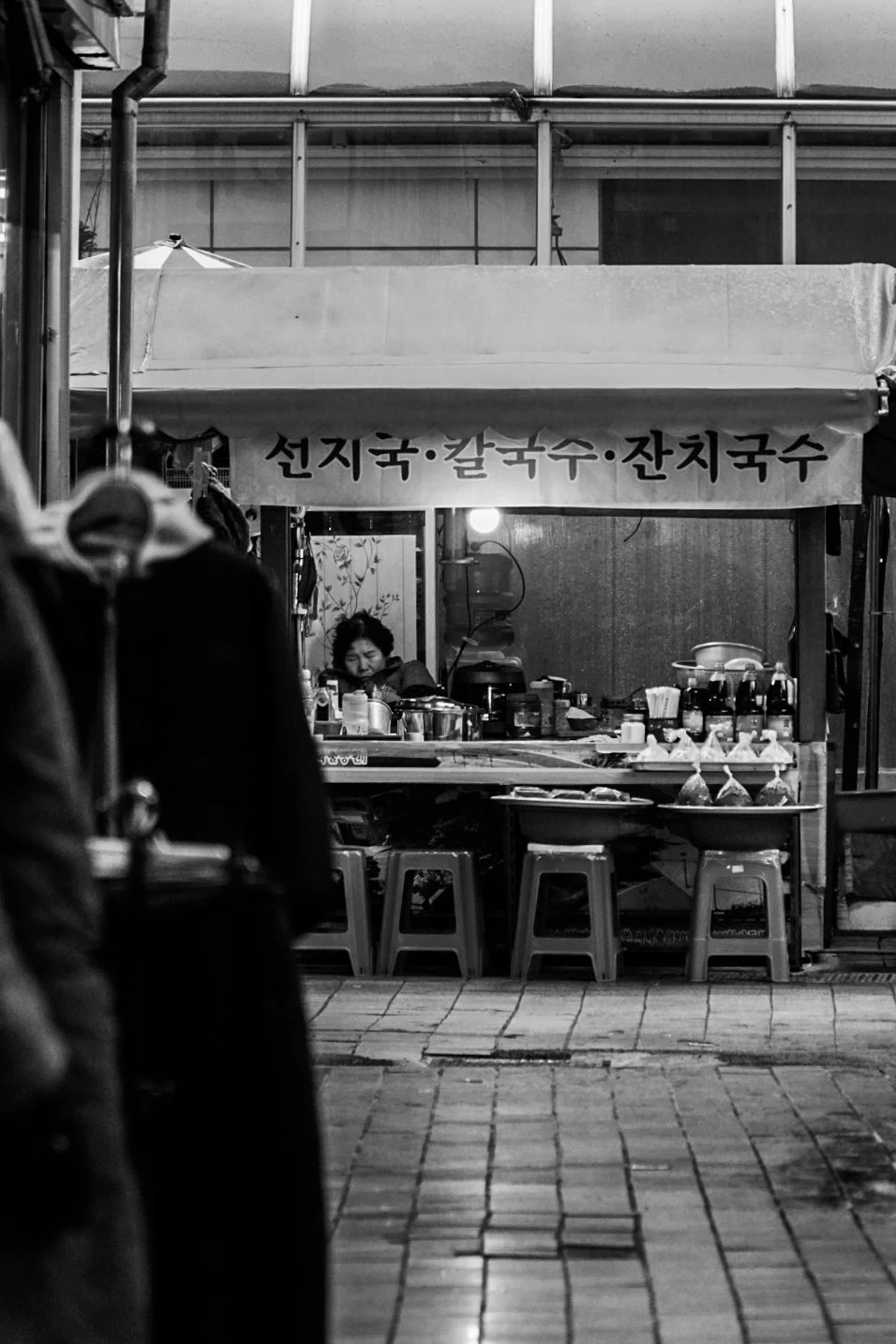 Street food stall with a vendor preparing food, Korean signs on a banner, stools and various food containers on the counter, night scene in black and white.