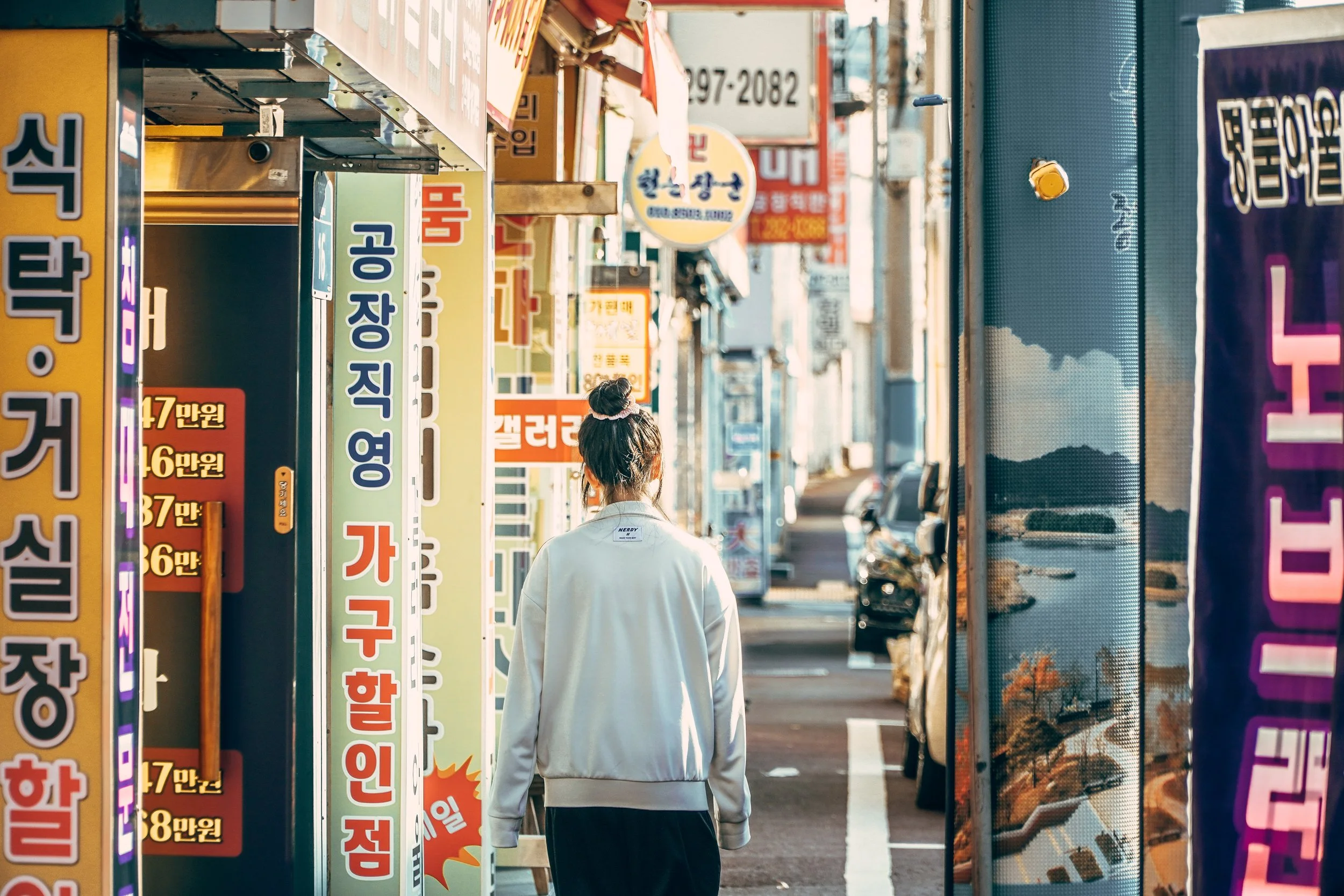 A person with tied-up hair, wearing a white sweatshirt, walking down a street flanked by colorful signs with Korean writing in a busy urban area.