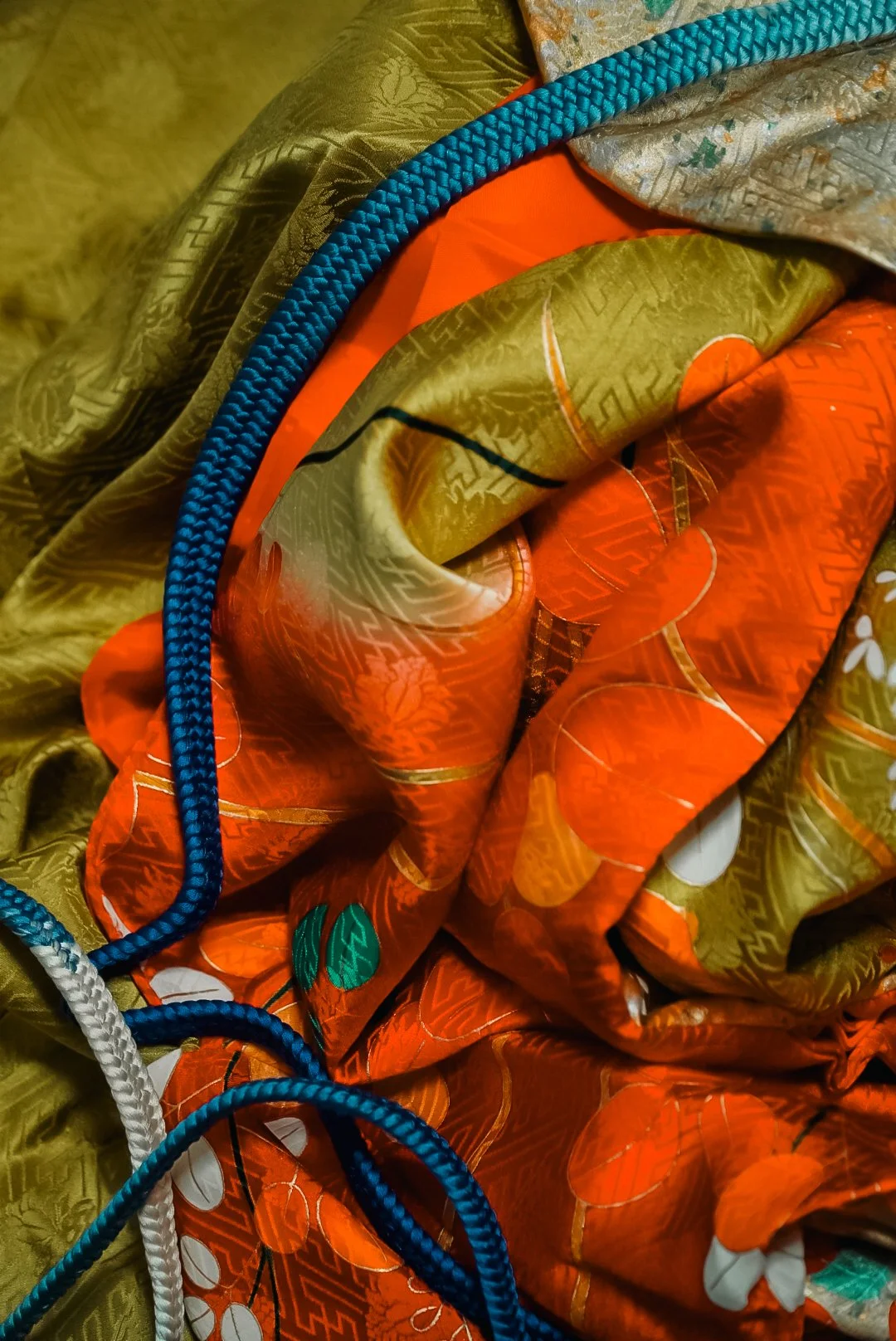 Close-up of brightly colored crumpled fabric with intricate patterns, featuring orange, gold, and white tones, alongside braided blue and white cords.
