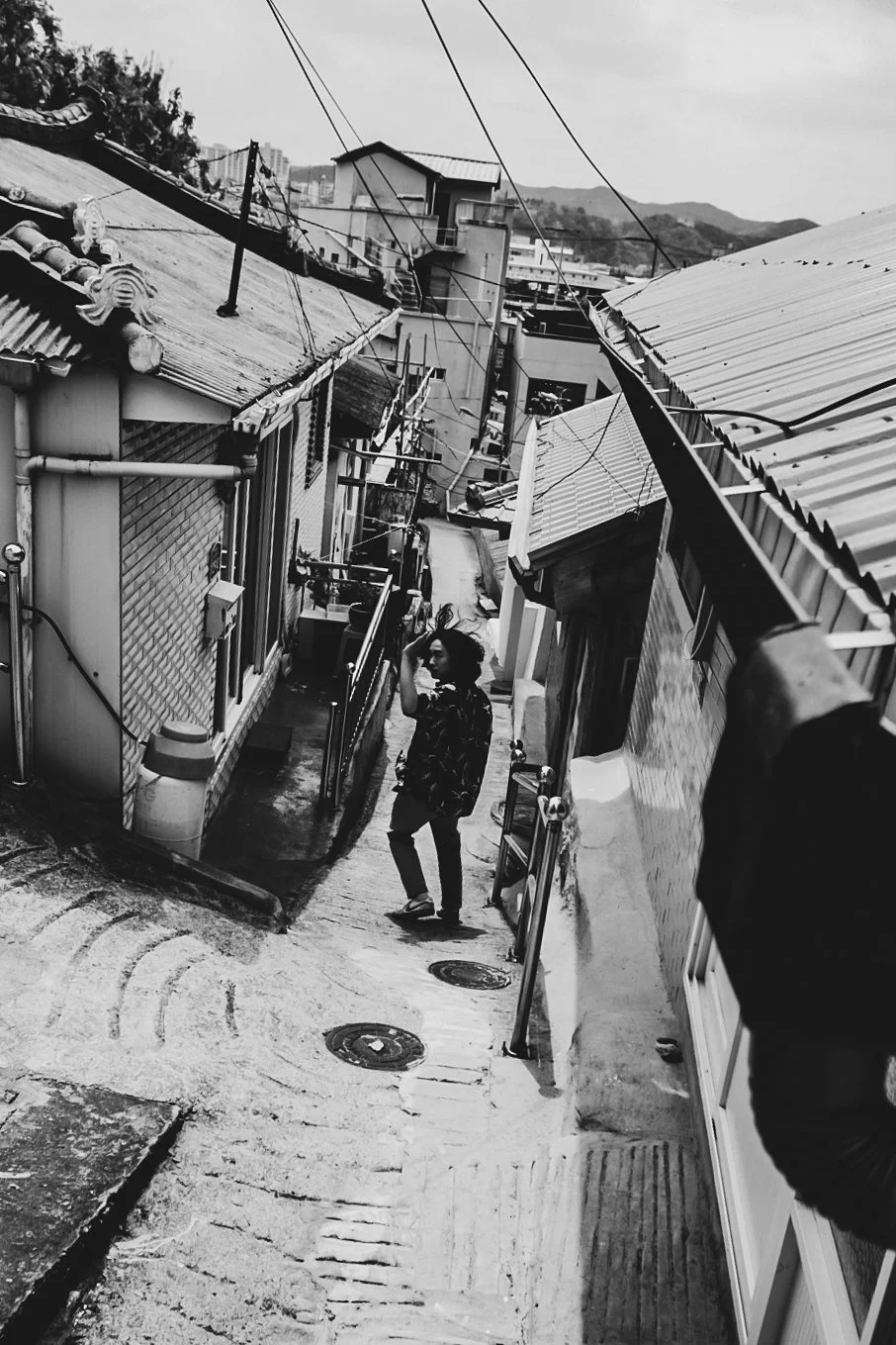 A woman standing on a sloped alleyway between closely packed houses with corrugated metal roofs, some visible utility equipment, and electrical wires overhead. She is smiling and appears to be waving.