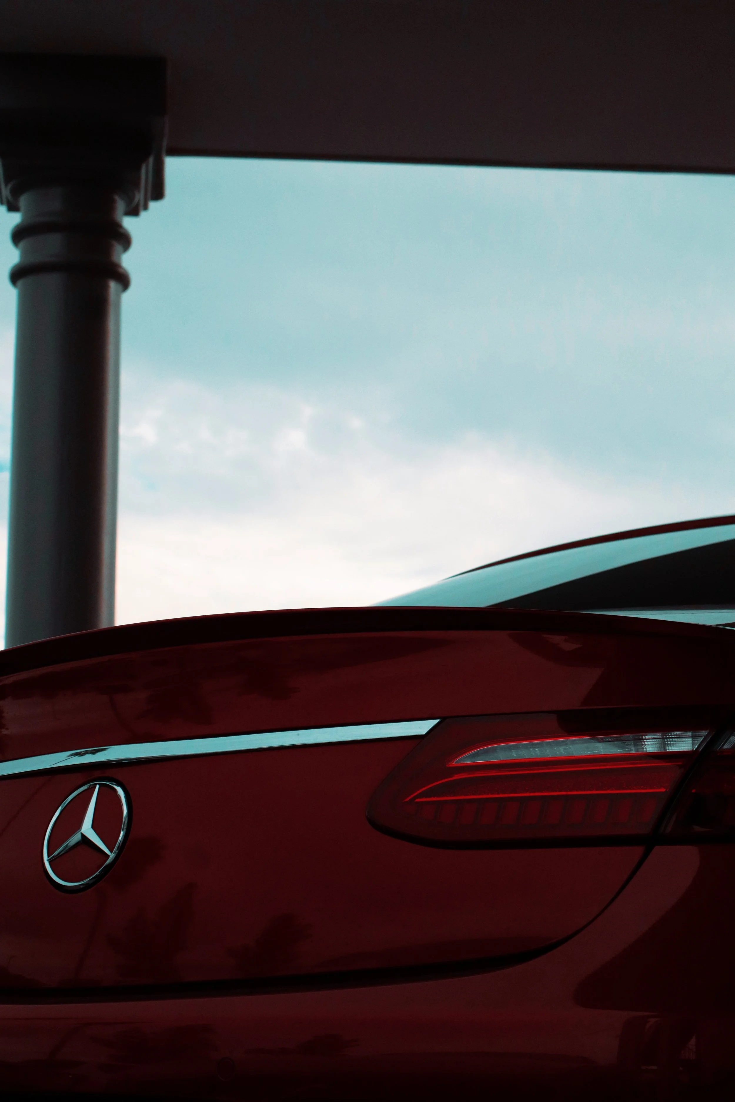 The rear end of a red Mercedes-Benz car showing its tail light and emblem, with the view of a blue sky and part of a metallic pole in the background.