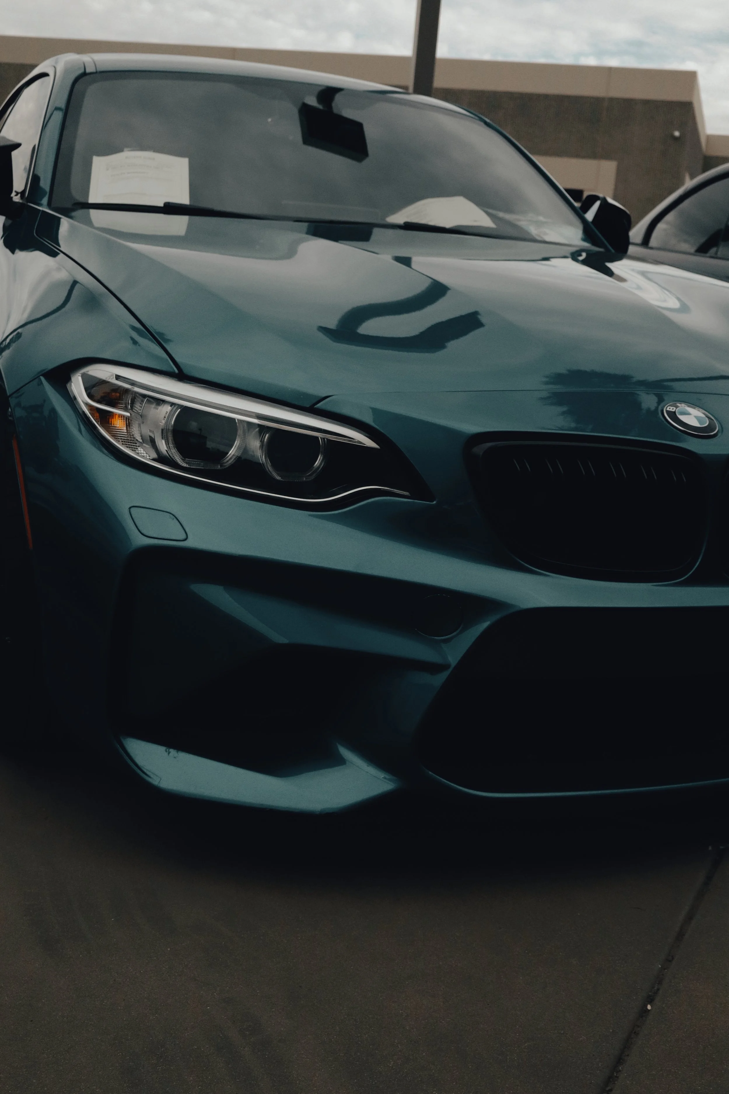 A blue BMW car parked outdoors with a white paper on the passenger side window and a black machine or device on the dashboard inside visible through the windshield.