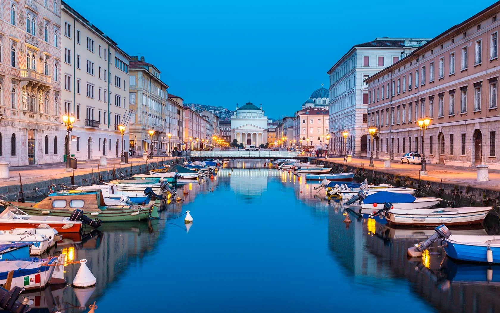 A city canal lined with boats and historic buildings, with a classical building and cathedral dome in the background, illuminated by streetlights at dusk.