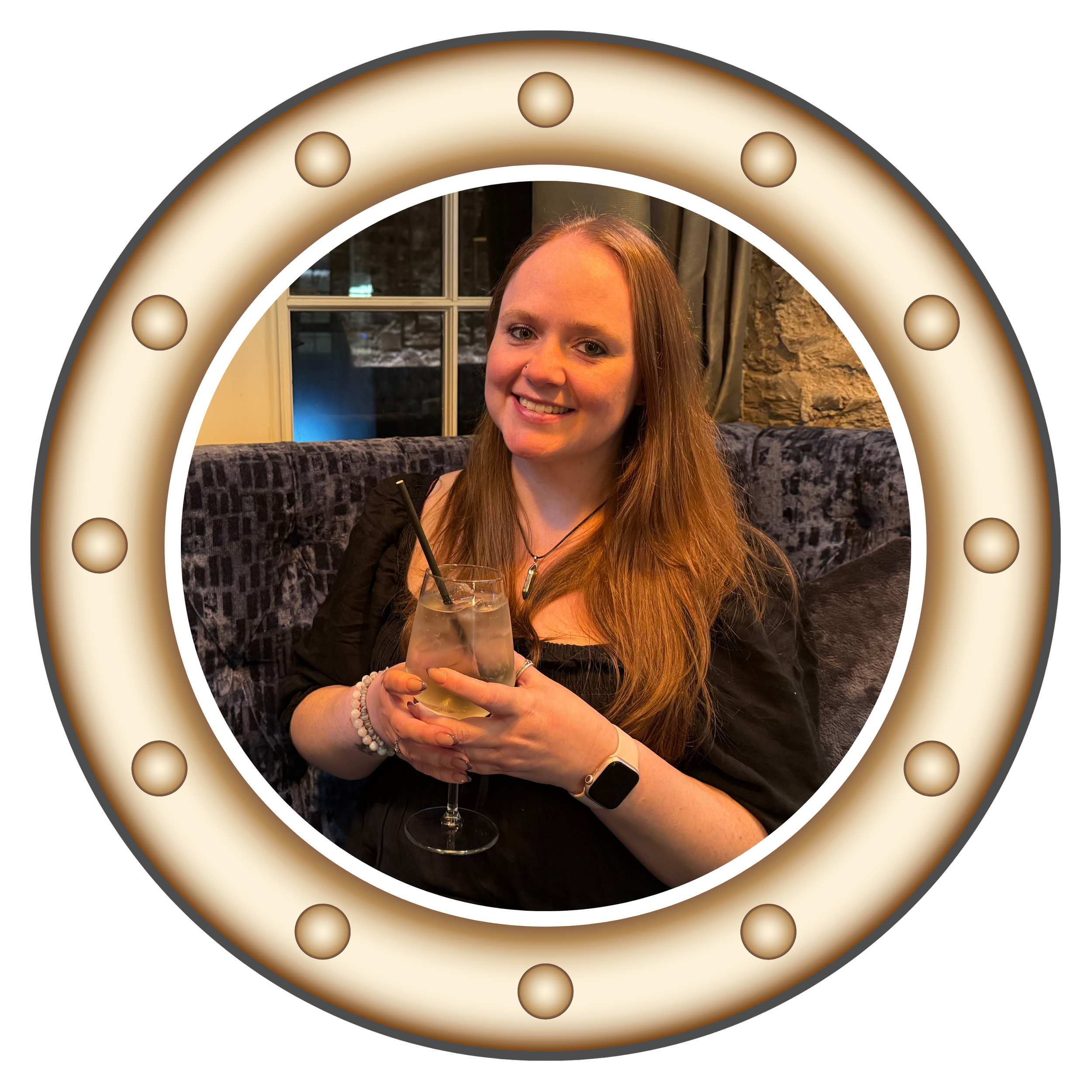 A woman with long hair and a nose piercing smiling at the camera while holding a glass of a clear drink with ice and a black straw, sitting on a black and grey patterned couch in a cozy room with a window and curtains.