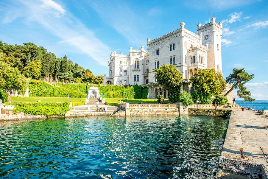 A large white castle-like building on a lakeshore, with lush green trees and gardens in front, calm blue water, and a partly cloudy sky.