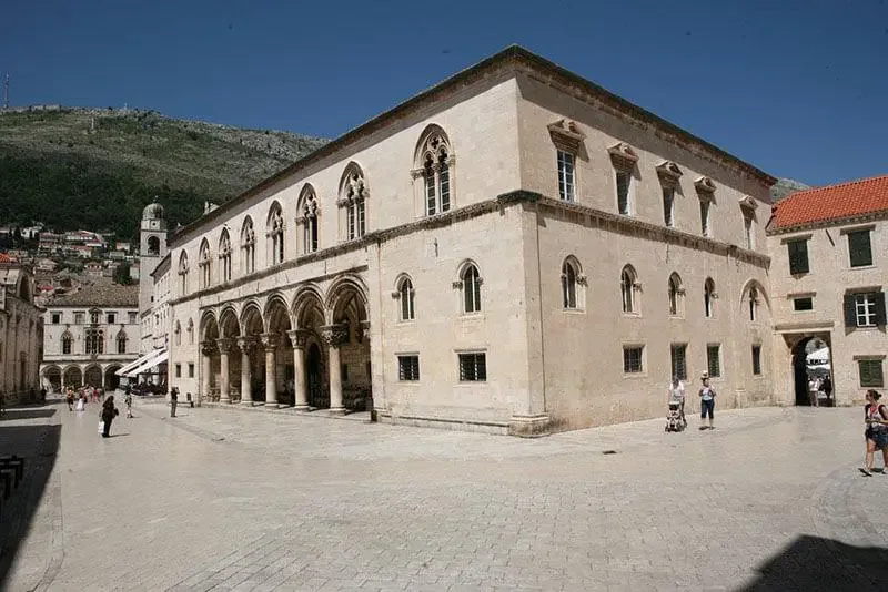 Historic stone building with arched windows and columns in a town square with people walking, backed by a hilly landscape under a clear blue sky.