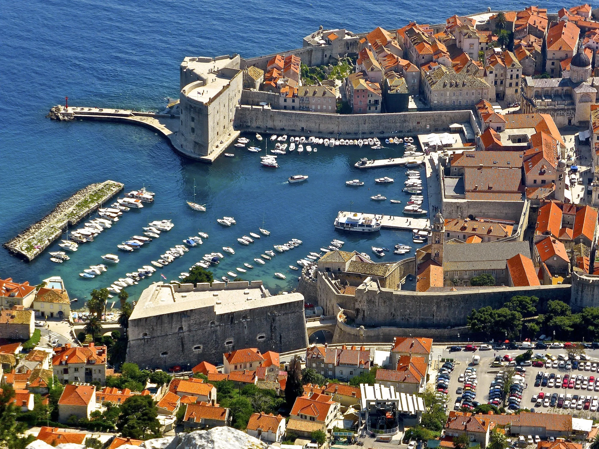 An aerial view of a historic fortified city by the sea with stone walls, red-tiled roofs, and a harbor filled with boats and yachts.