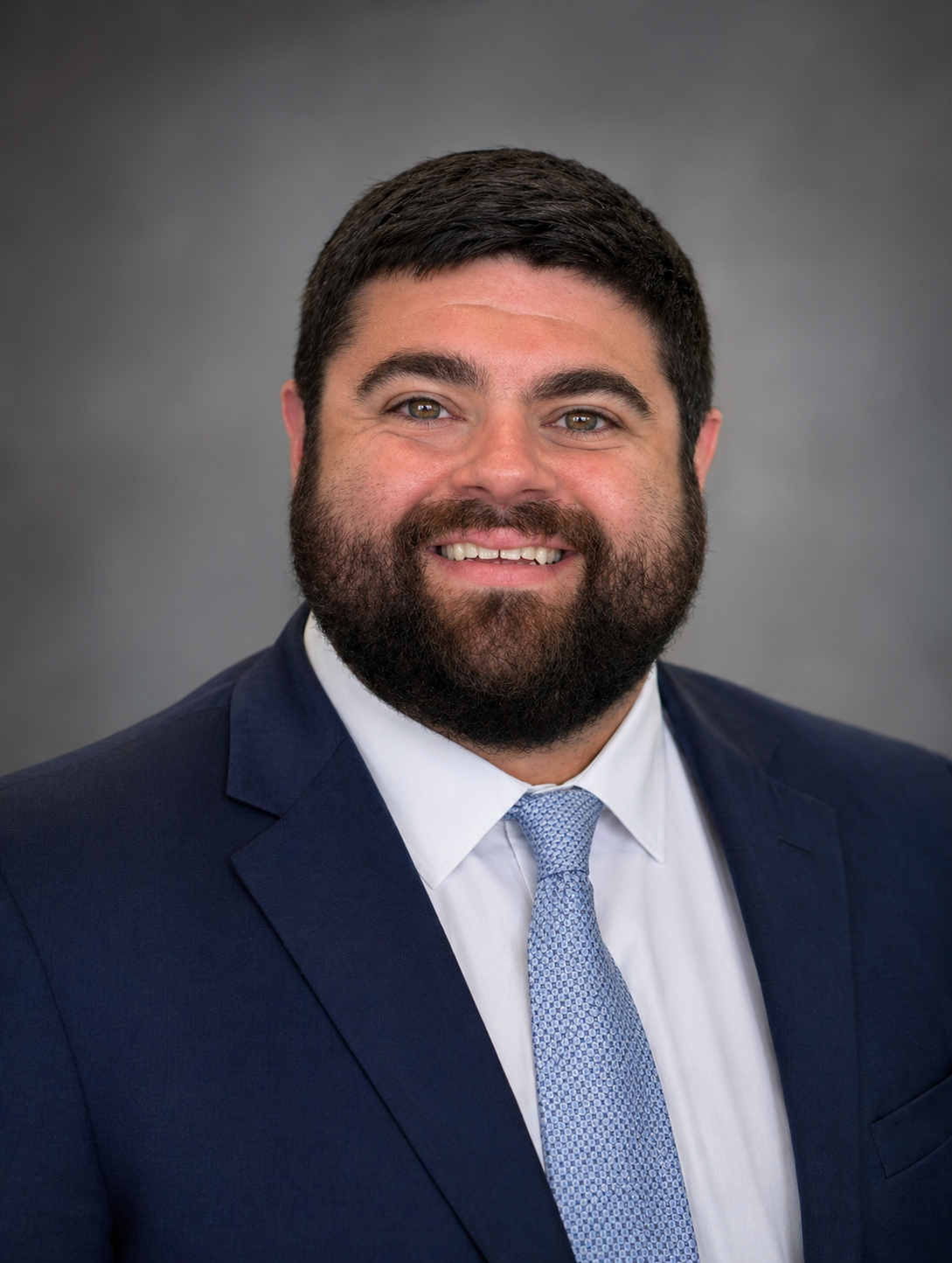 Headshot of a man with dark hair and beard wearing a navy suit, white shirt, and light blue tie, smiling against a gray background.