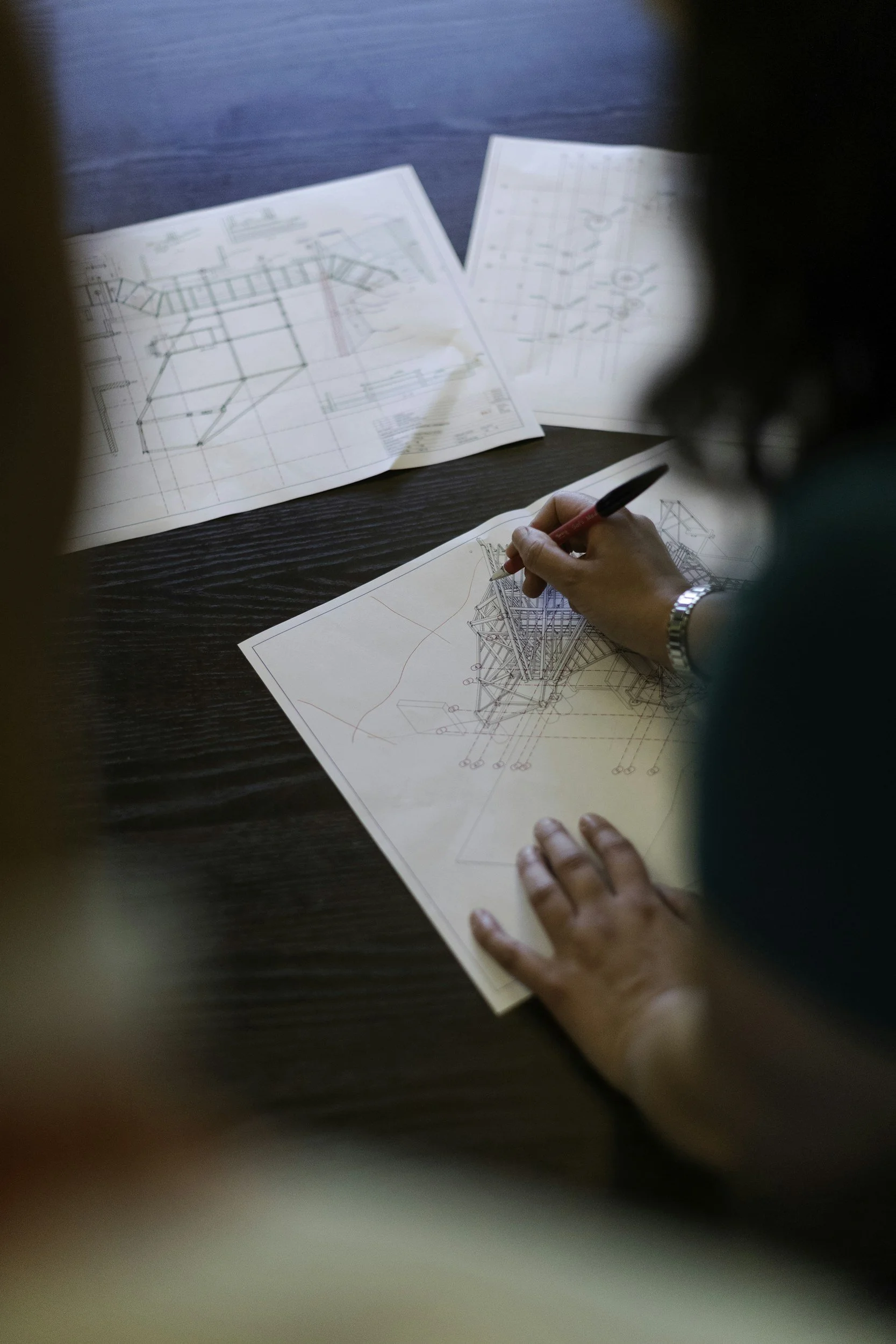 To personer, der arbejder med arkitekttegninger og planer på et bord, med fokus på en persons hånd, der holder en sort og rød kuglepen, og ark med bygningstegninger.