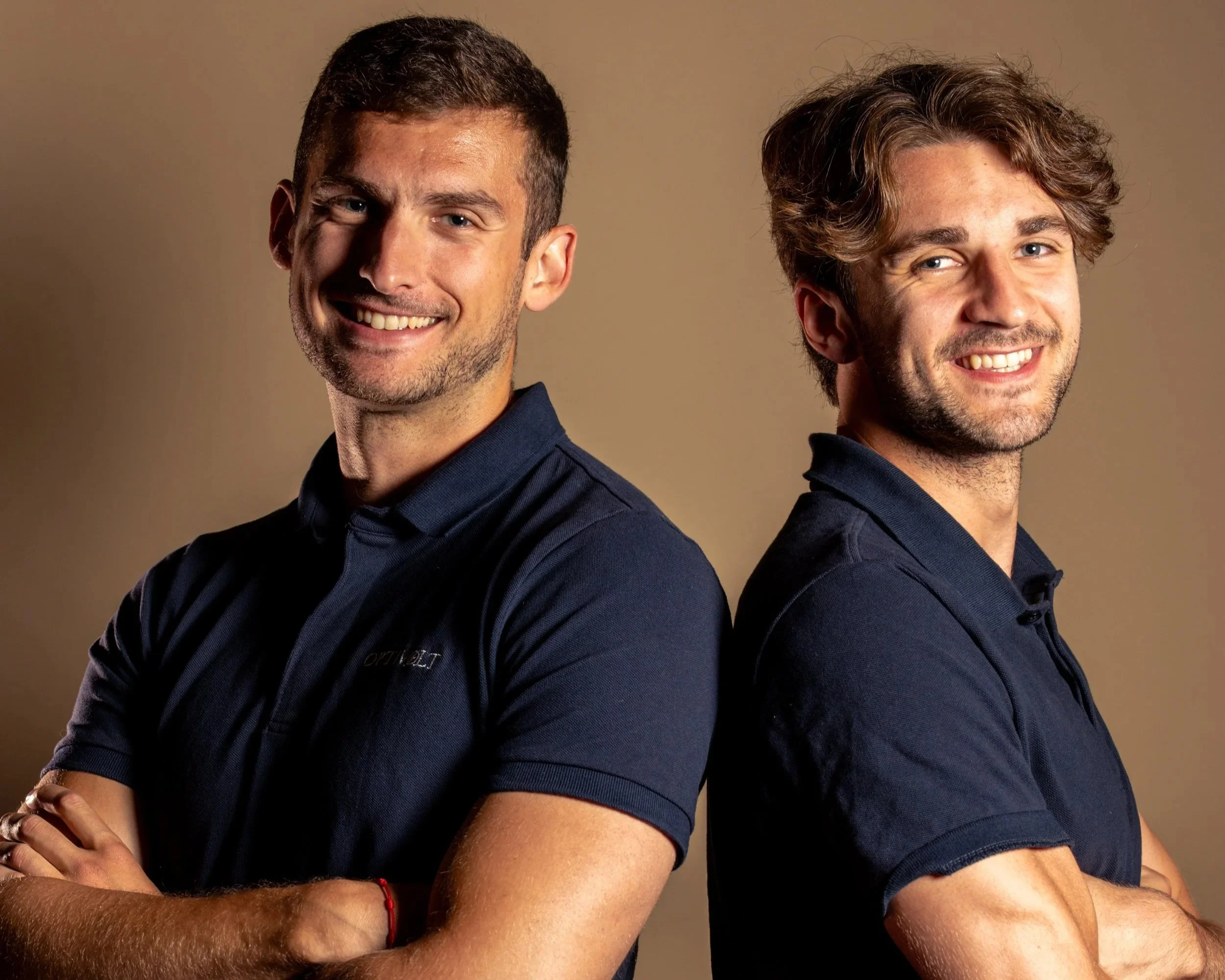 Two young men smiling, standing back to back, with arms crossed, against a plain beige background. They are wearing dark blue shirts.