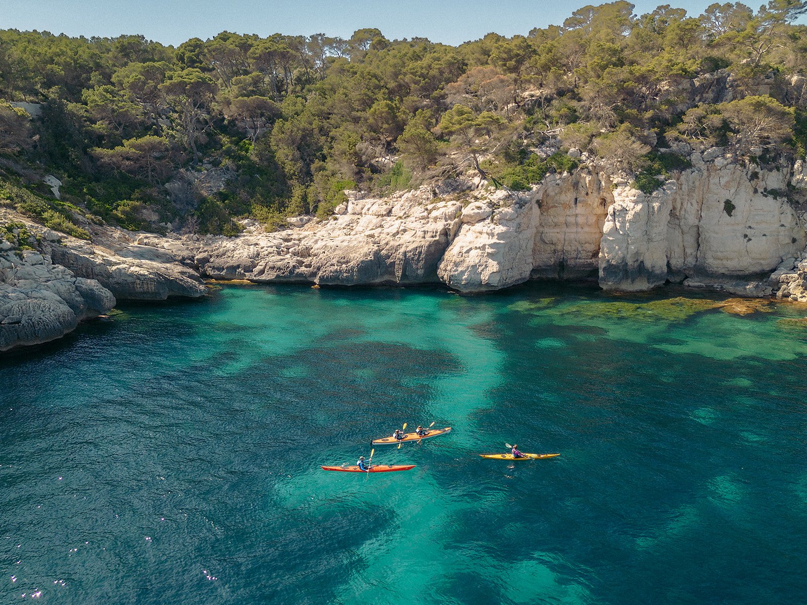 Varios kayaks de mar recorren una cala de aguas cristalinas en Menorca, rodeada de acantilados y vegetación mediterránea