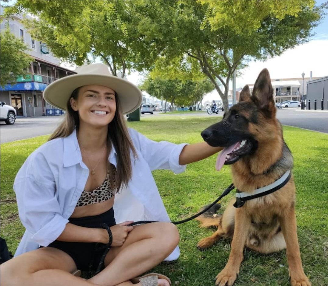 A young woman smiling while sitting on the grass in a park with her German Shepherd dog, under a large leafy tree with a city street, parked cars, and buildings in the background.