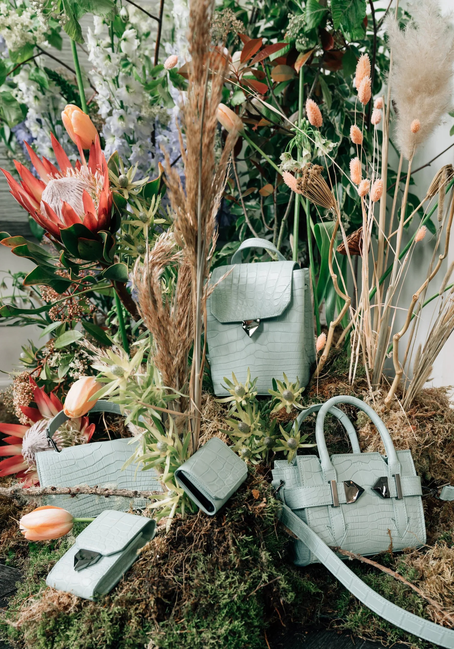 Shwaz sage-green crocodile-leather handbags with gold hardware, styled among protea and dried grasses.