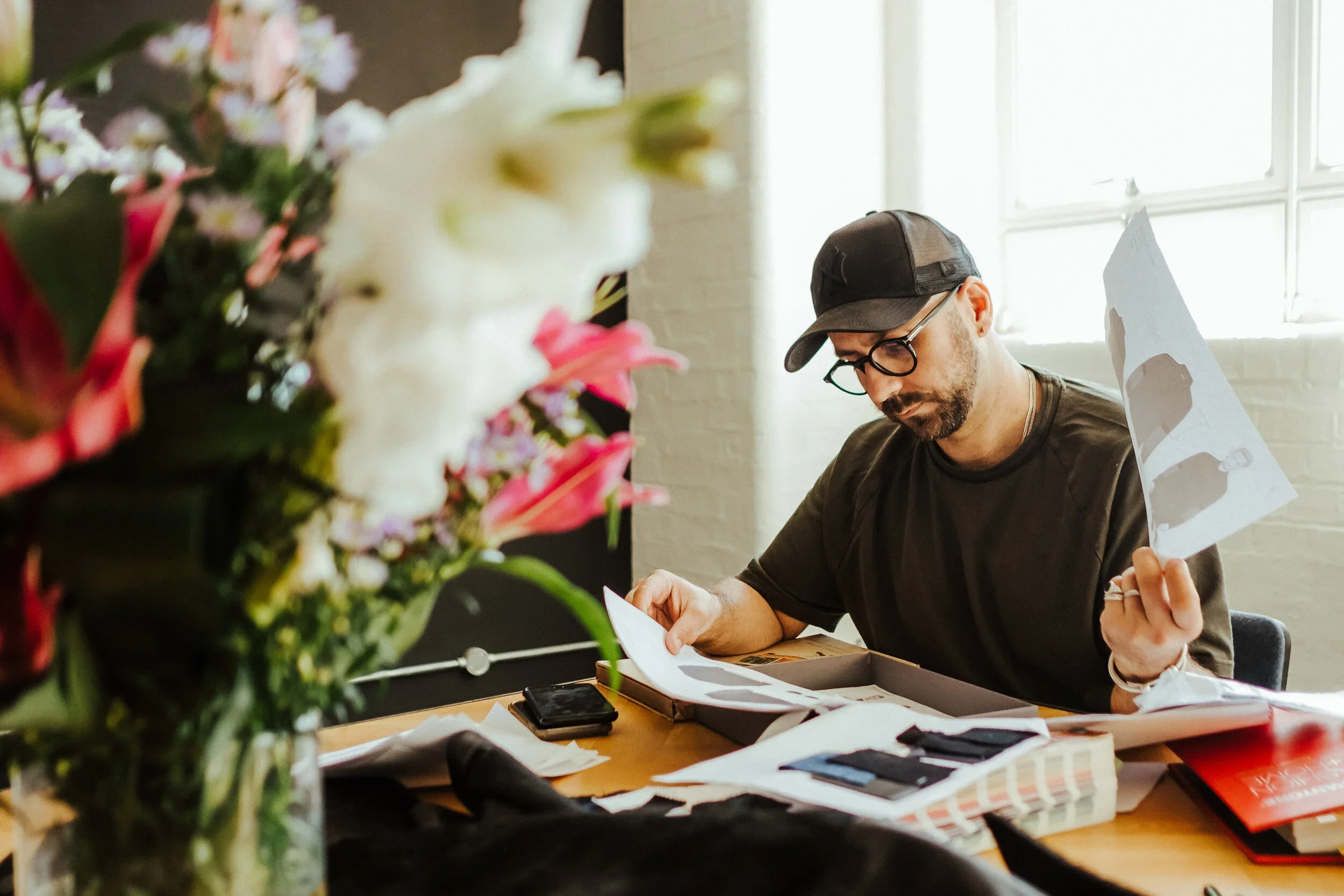 A man with glasses, a beard, wearing a black cap and black t-shirt, sitting at a cluttered table reviewing papers.