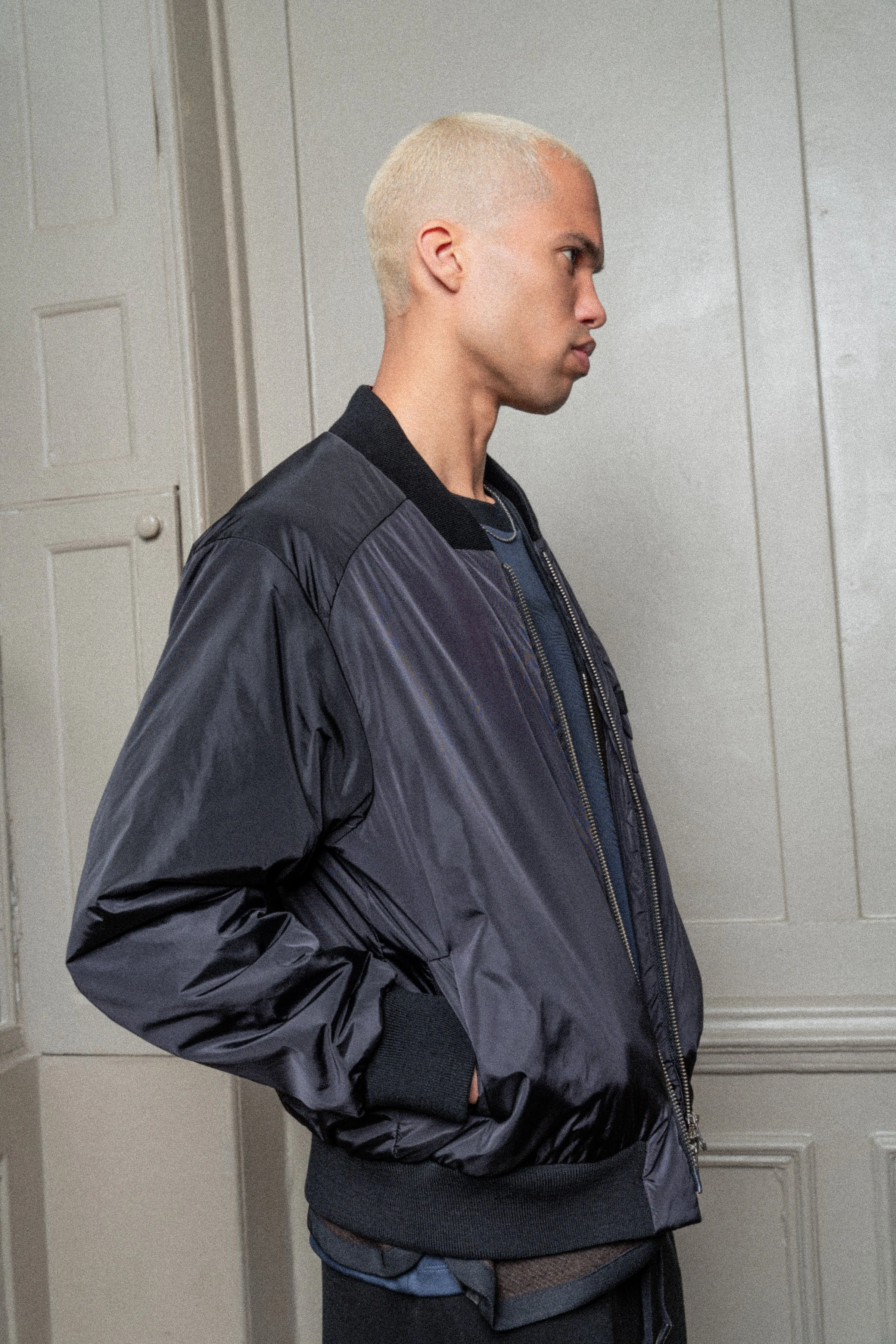 Side view of a young man with short platinum blonde hair, wearing a black and gray satin bomber jacket and standing with hands in pockets, against a white paneled wall.