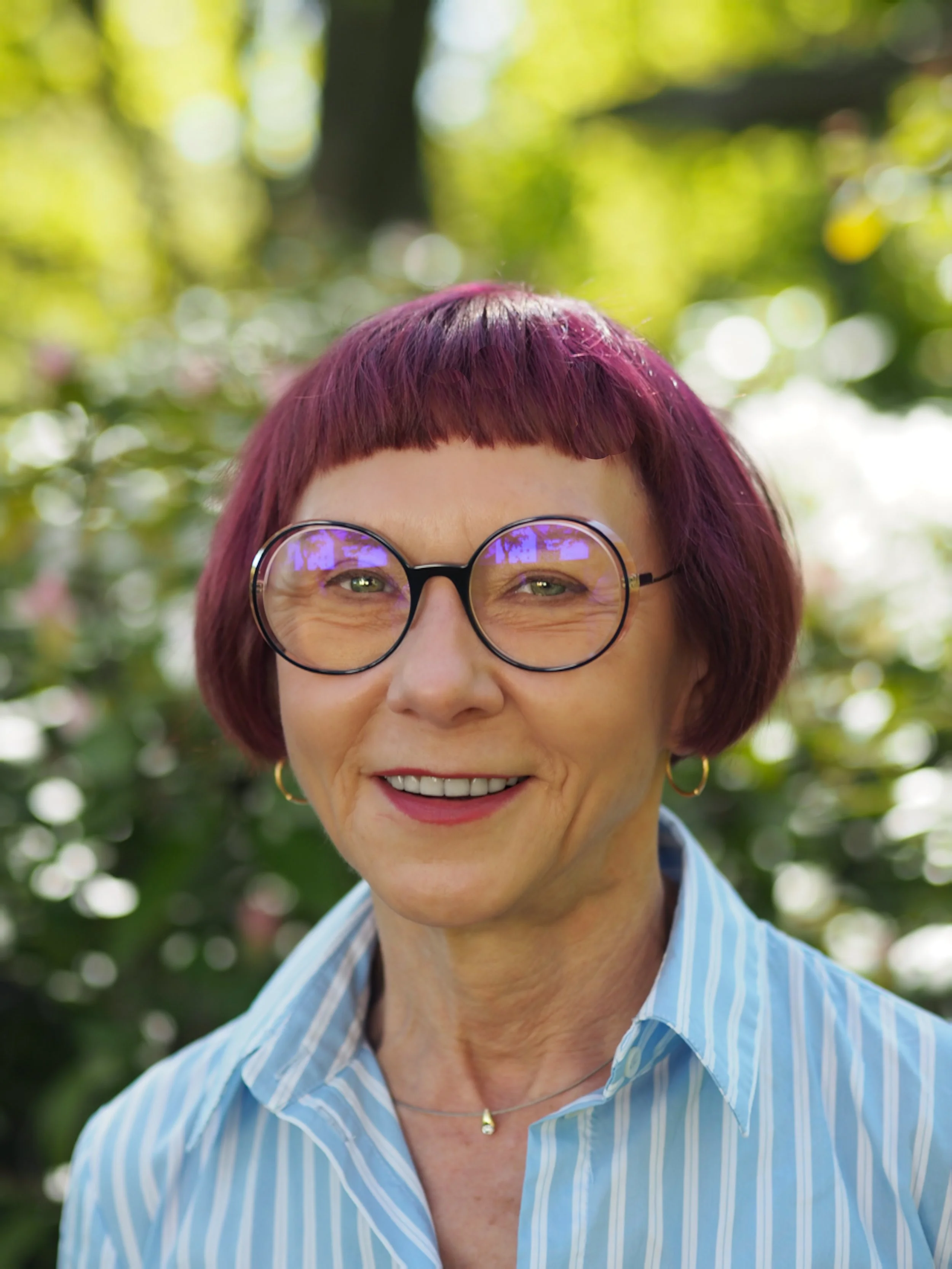 Nahaufnahme eines lachenden Frauenkopfes mit roten Haaren, runden Brillen und einem blauen gestreiften Hemd, im Hintergrund grüner Garten mit Bokeh-Effekt.