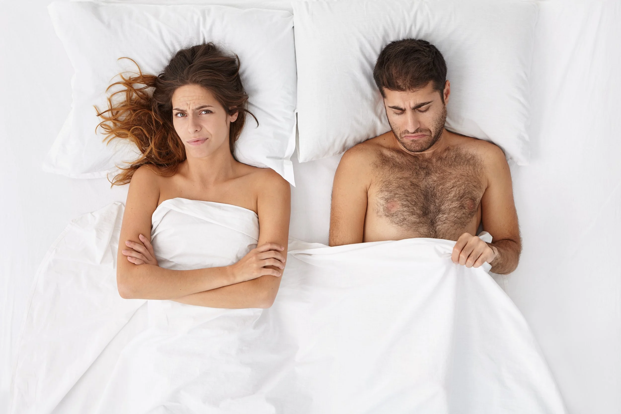 Ein Mann und eine Frau liegen nebeneinander in einem Bett, beide haben eine skeptische oder nachdenkliche Miene. Der Mann ist nackt bis auf den Oberkörper, die Frau ist mit einem weißen Tuch bedeckt.