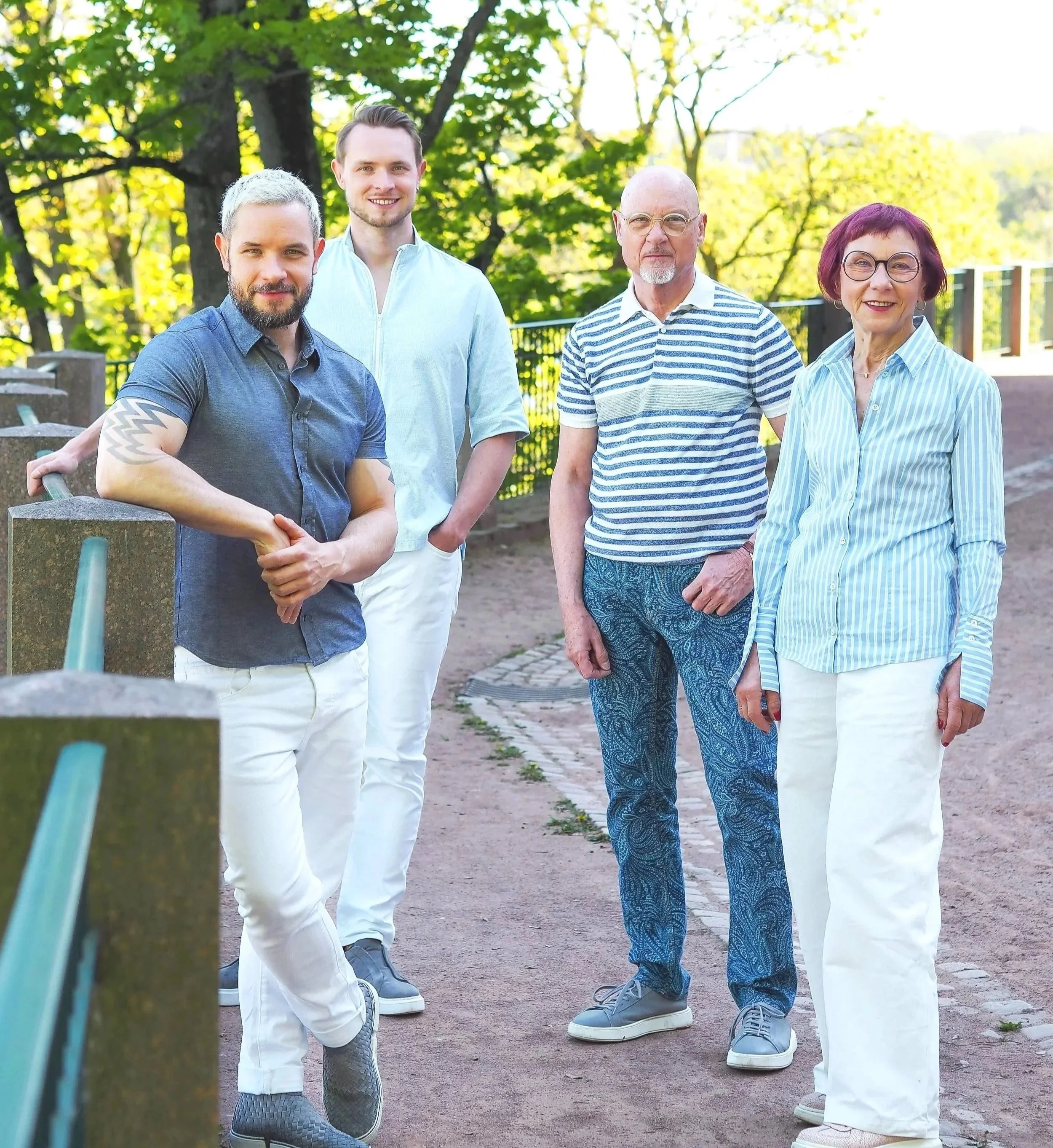Fünf Personen stehen im Freien auf einem Weg neben einem Zaun, umgeben von grünen Bäumen, und schauen in die Kamera. Es ist Tag, sie tragen legere Kleidung. Zwei Männer haben graues und braunes Haar, eine Frau hat rotbraune Haare und trägt größere Brillen, ein älterer Mann hat eine Glatze und trägt eine Brille.