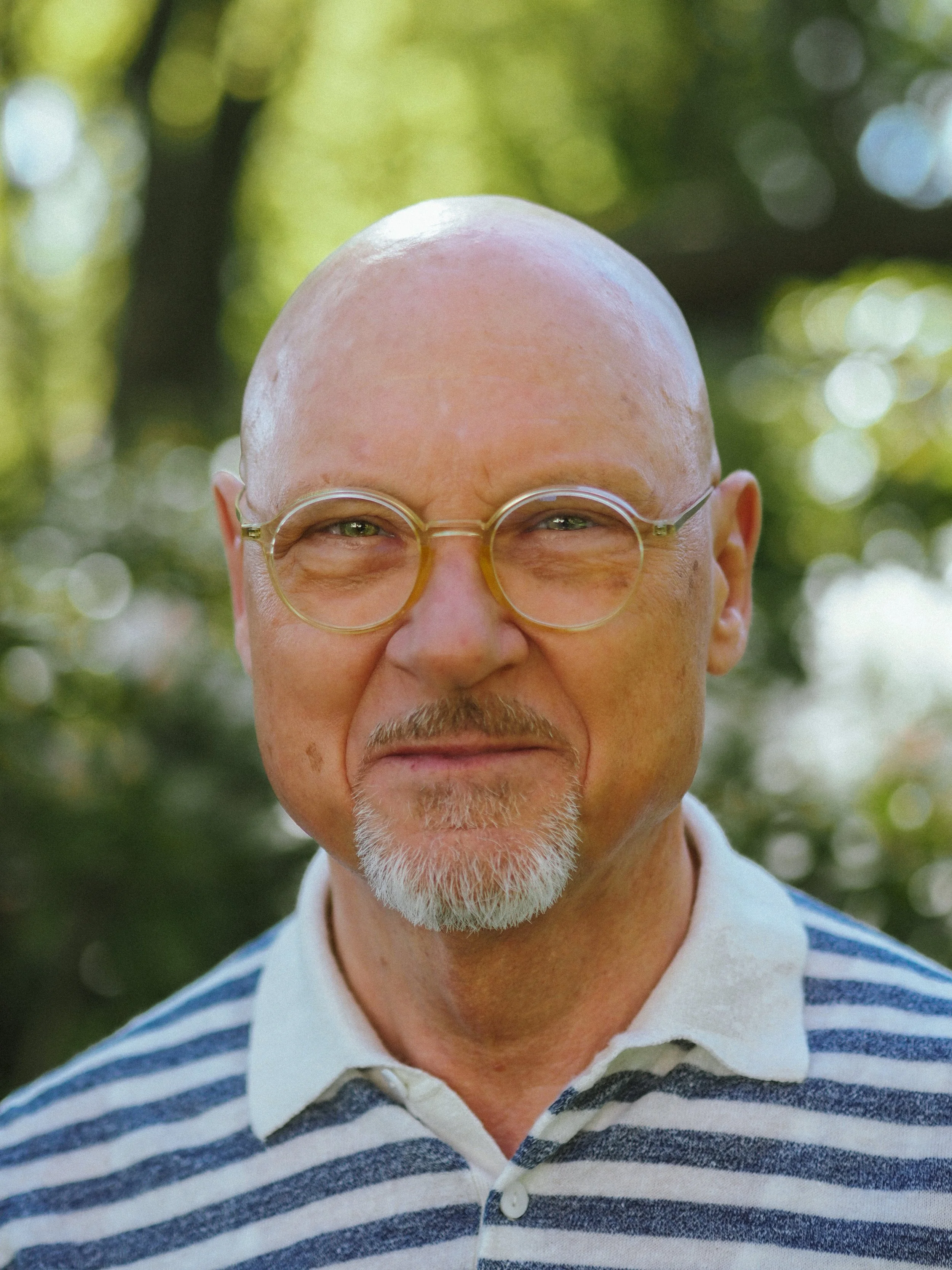 Nahaufnahme eines älteren Mannes mit Glatze, Brille und weißem Bart, trägtdurchschaubare, runde Brille, trägt ein weiß-blau gestreiftes Hemd, im Freien, unscharfer grüner Hintergrund