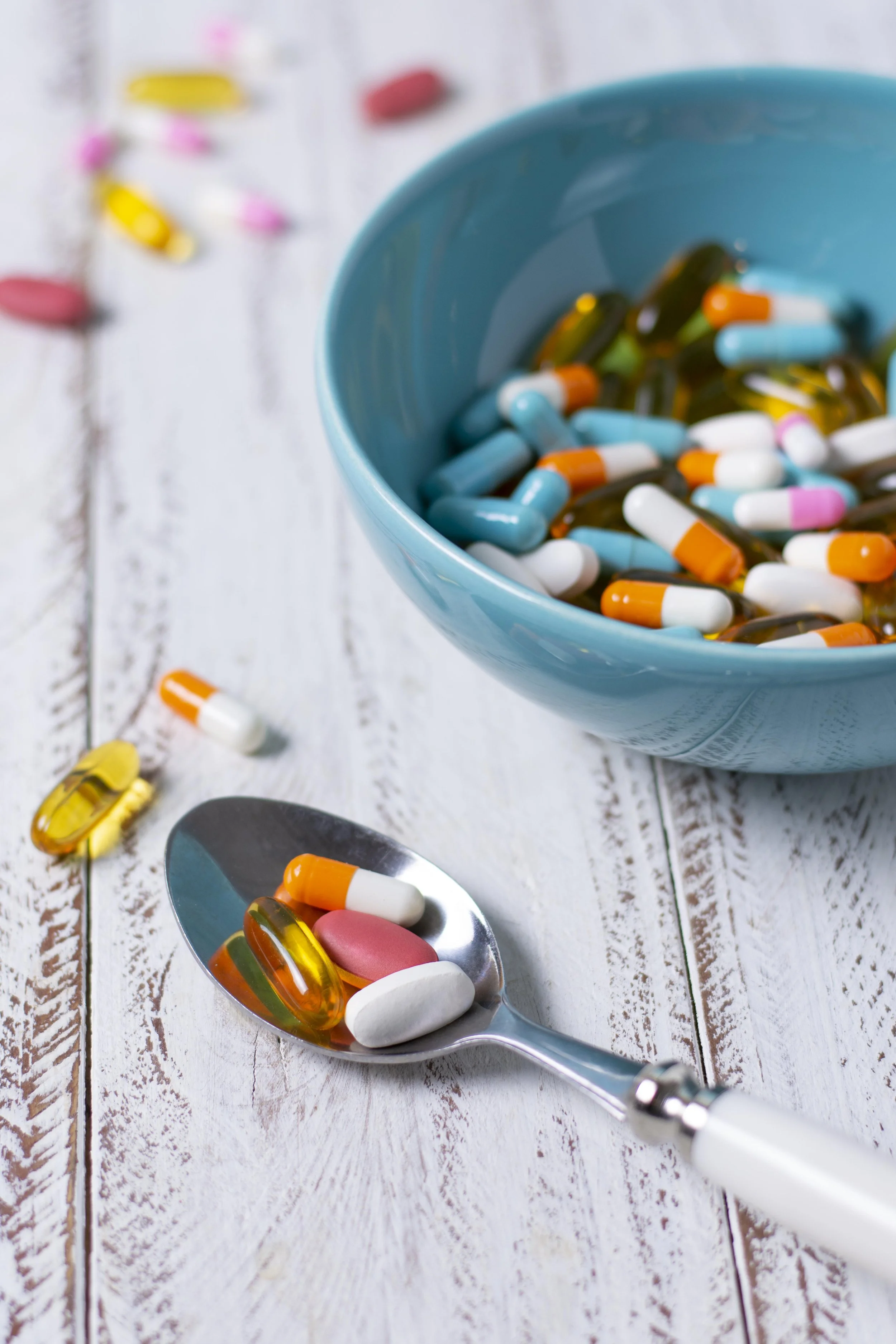 Bunte Kapseln und Tabletten in einer blauen Schüssel und auf einem Löffel, auf einem weißen Holztisch