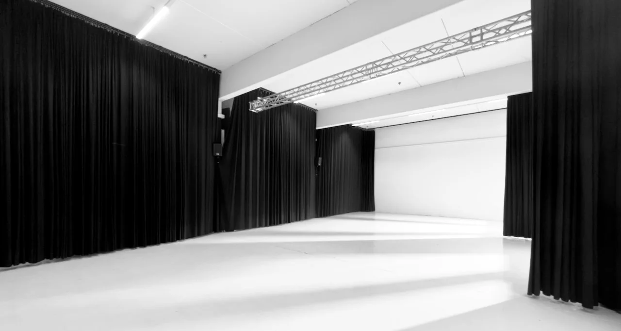 Schwarz-weißer Veranstaltungsraum mit schwarzen Bühnenvorhängen, weißen Wänden, schwarzem Bühnenvorhang, weißem Boden und Deckenbeleuchtung.
