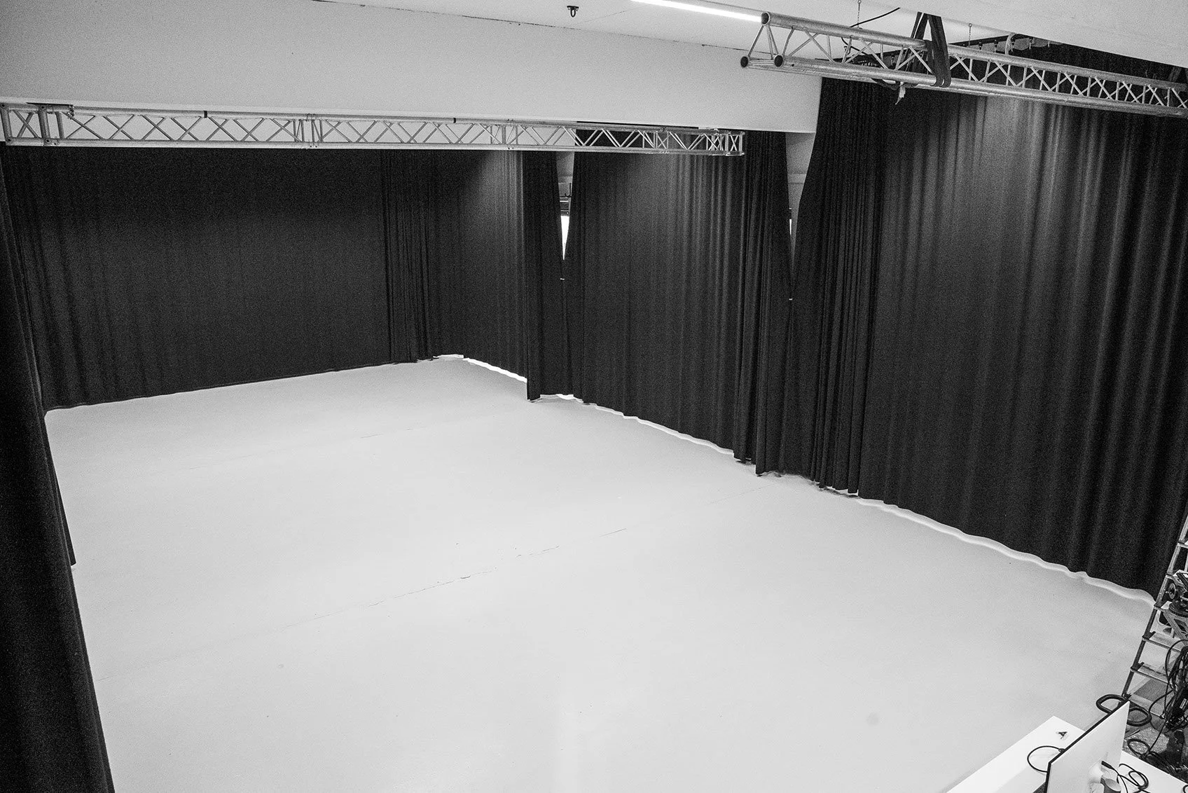 Ein leeres Bühnen- oder Studiozimmer mit schwarzen Vorhängen, einer weißen Bodenfläche und einer technischen Leiter an der rechten Seite.