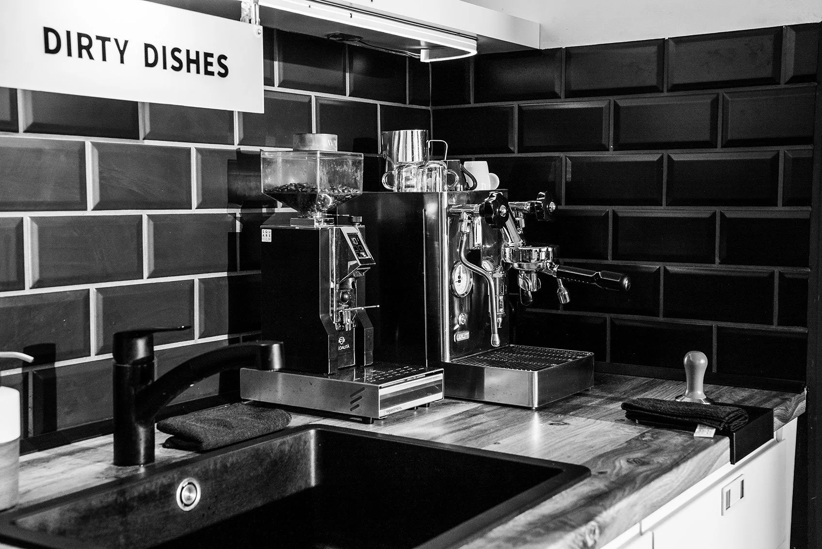Eine Kaffeemaschine und eine Kaffeemühle stehen auf einer Holztheke vor einer schwarz glasierten Wand. Es gibt einen schwarzen Wasserhahn im Vordergrund und ein handgeschlagenes schwarzes Tuch oder eine Serviette auf der Theke.