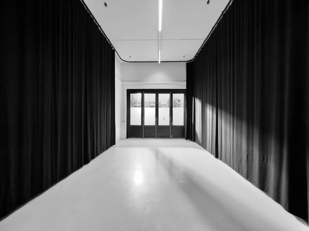 Ein leerer Raum mit schwarzen Vorhängen an den Seiten, einer weißen Bodenfläche und einem Fenster mit Türen am Ende, alles in Schwarz-Weiß.