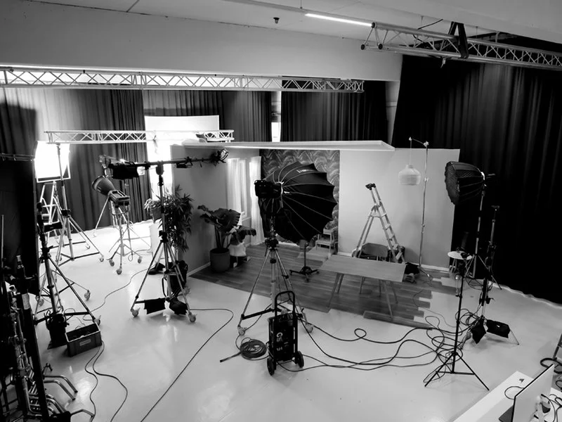Fotostudio mit professioneller Beleuchtung, Kameras, Stühlen, Tischen, einem Leiter, dunklen Vorhängen und technischen Geräten für Fotoshootings oder Videoaufnahmen.