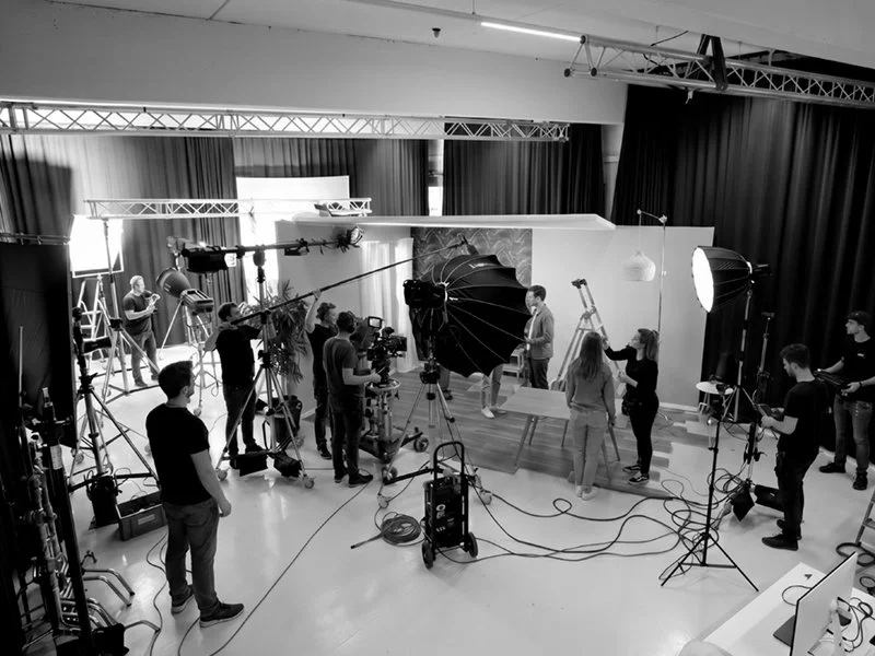 Innenaufnahme eines Film- oder Fotoshootings mit mehreren Personen, Kamera- und Beleuchtungsausrüstung, und einem Studio-Hintergrund.
