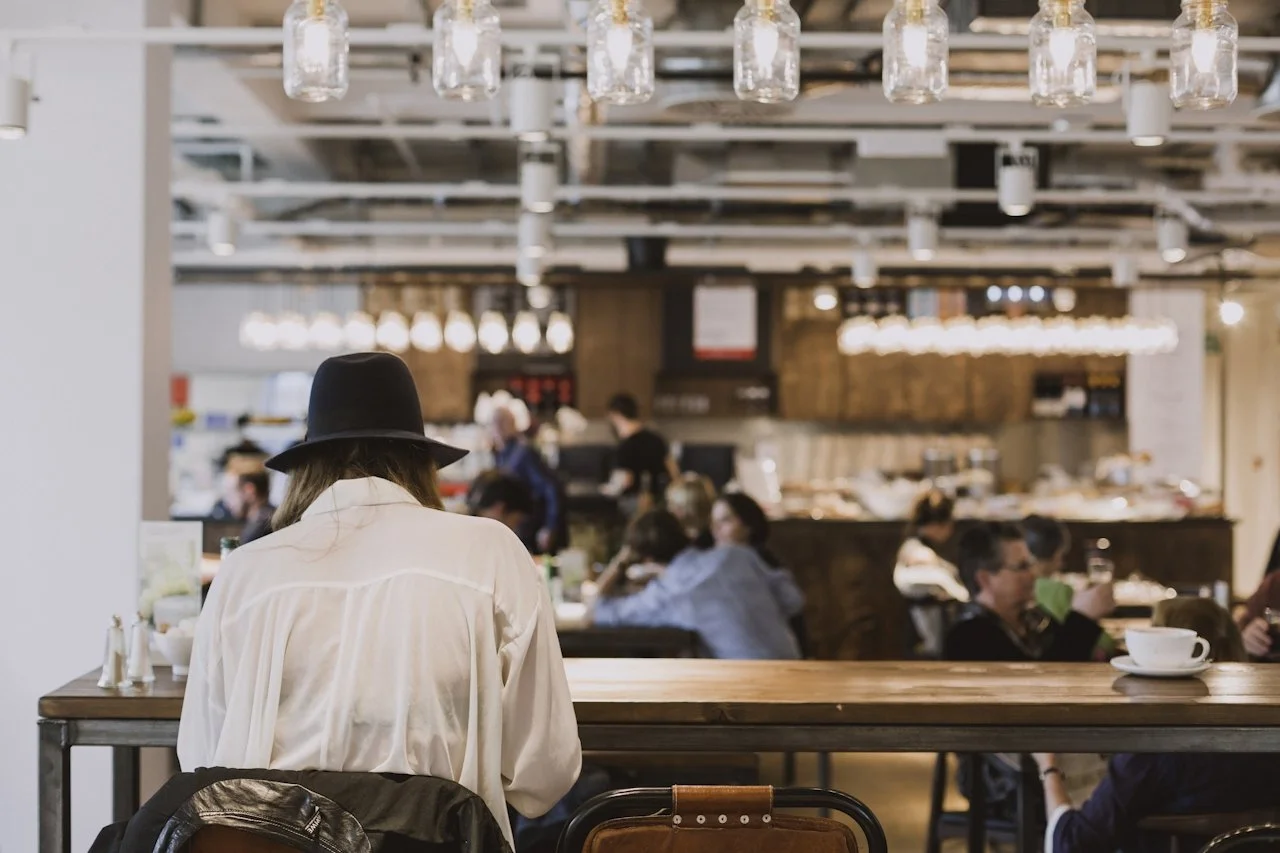 Woman sitting in cafe in white shirt and black hat with back towards camera