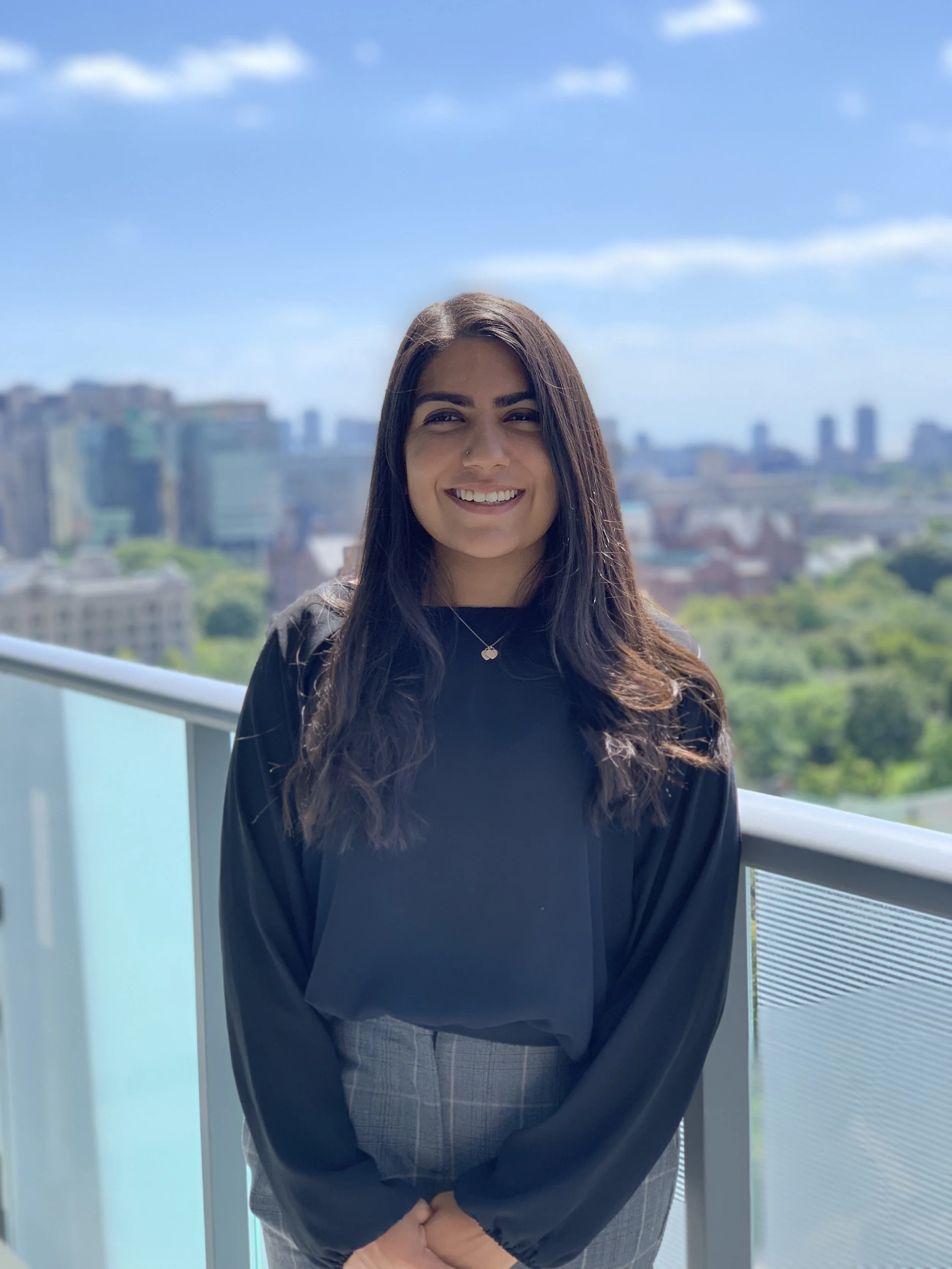 A young woman with long dark hair, wearing a black top and gray checked pants, stands outdoors on a balcony with city buildings in the background. She is smiling and looking at the camera, with a blue sky and some clouds above.