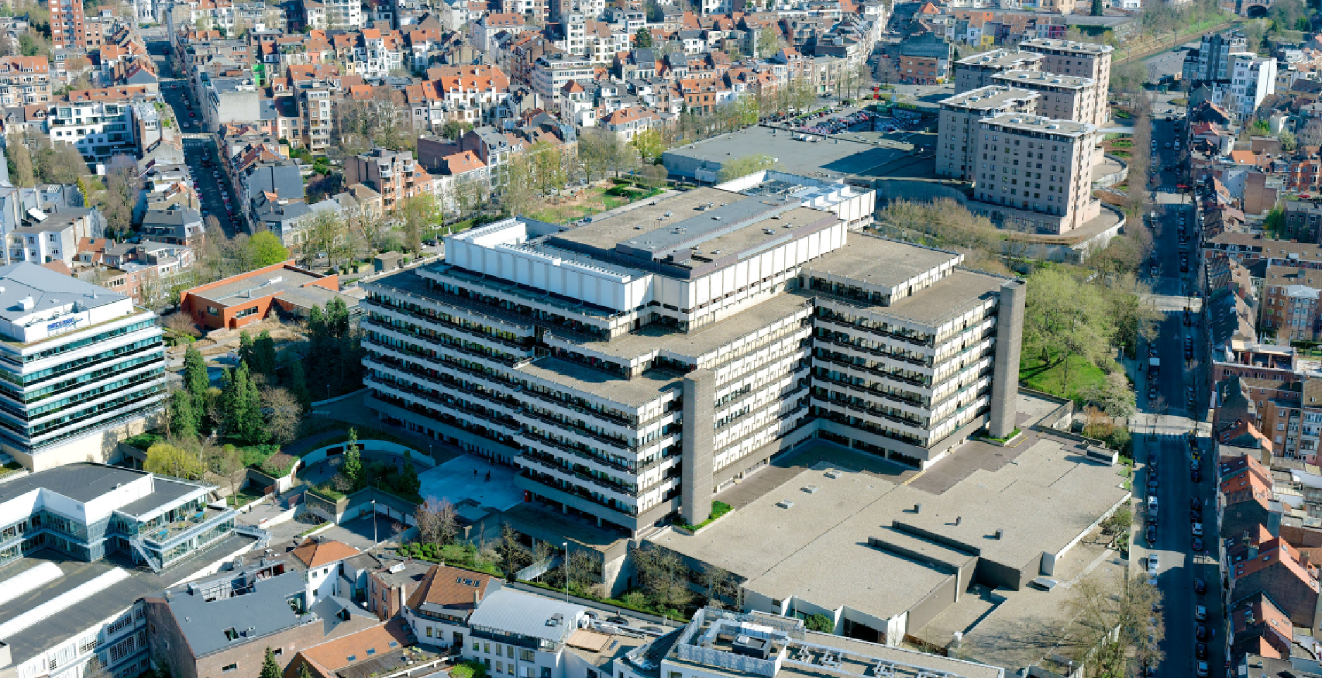 Vue aérienne d'une grande ville avec un bâtiment moderne de plusieurs étages au centre, entouré de quartiers résidentiels et de routes. La zone est verdoyante avec de nombreux arbres.