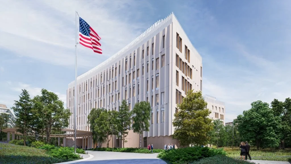 Un bâtiment moderne blanc avec forme géométrique, entouré d'arbres et avec un drapeau américain flottant au vent, sous un ciel bleu avec quelques nuages.