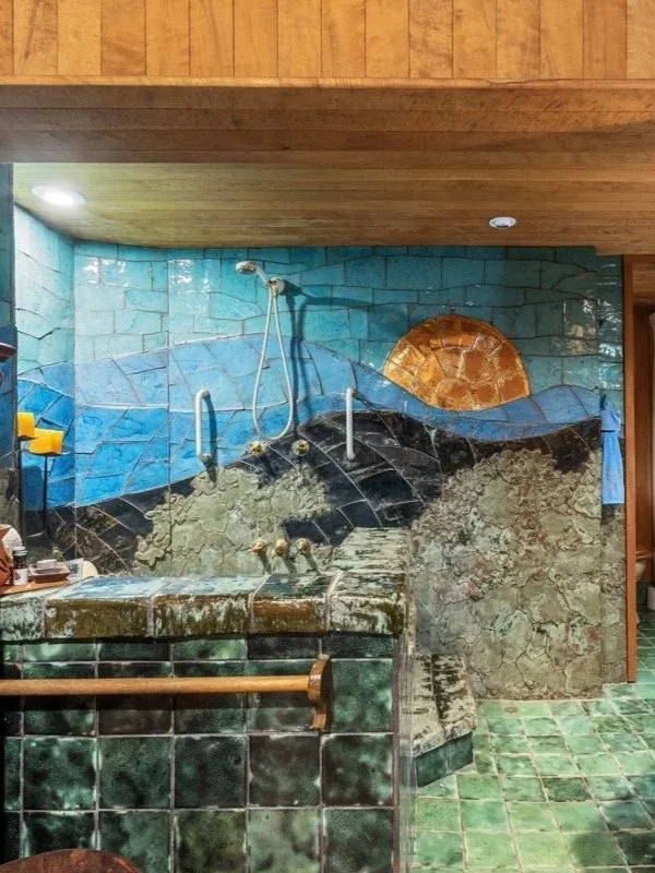 Decorative tiled wall with a mountain landscape mural, featuring a sun, blue sky, and green ground, in a spa or bathhouse setting.