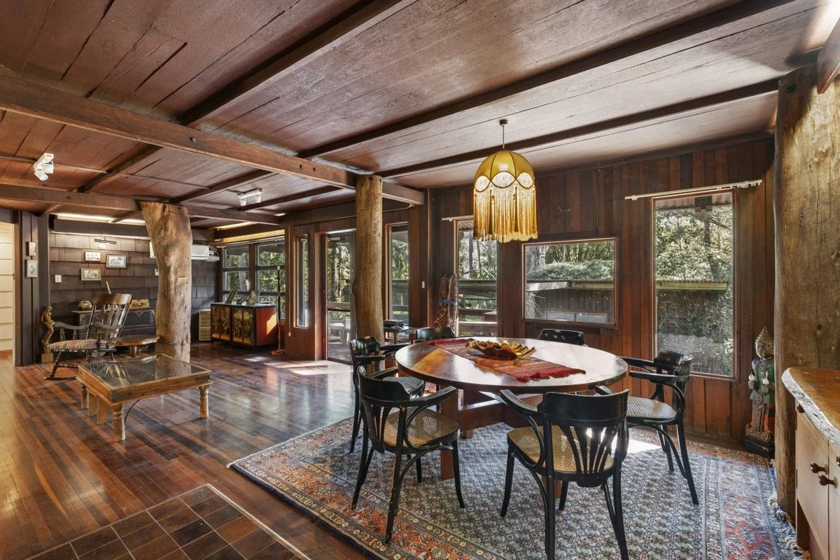 Interior view of a rustic wooden dining room with a round table, six black chairs, wood-paneled walls, large windows, and a vintage hanging lamp.